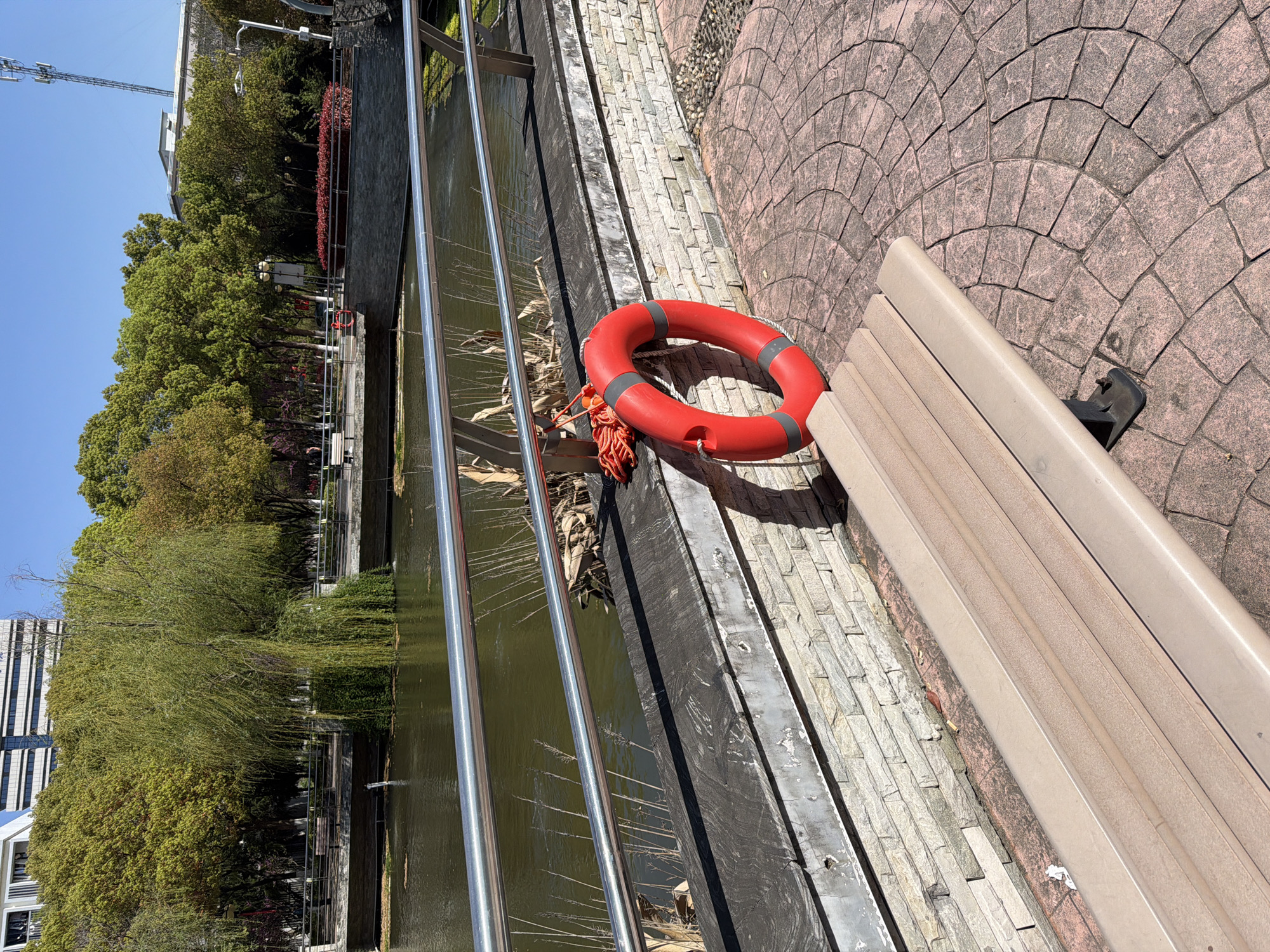 A red life preserver rests on a park bench beside a canal in Jiangning District, China. The canal is bordered by a metal railing and stone walls, with trees and foliage visible along the opposite bank. The bench sits on a paved walkway with a circular brick pattern. The scene appears to be a sunny day.