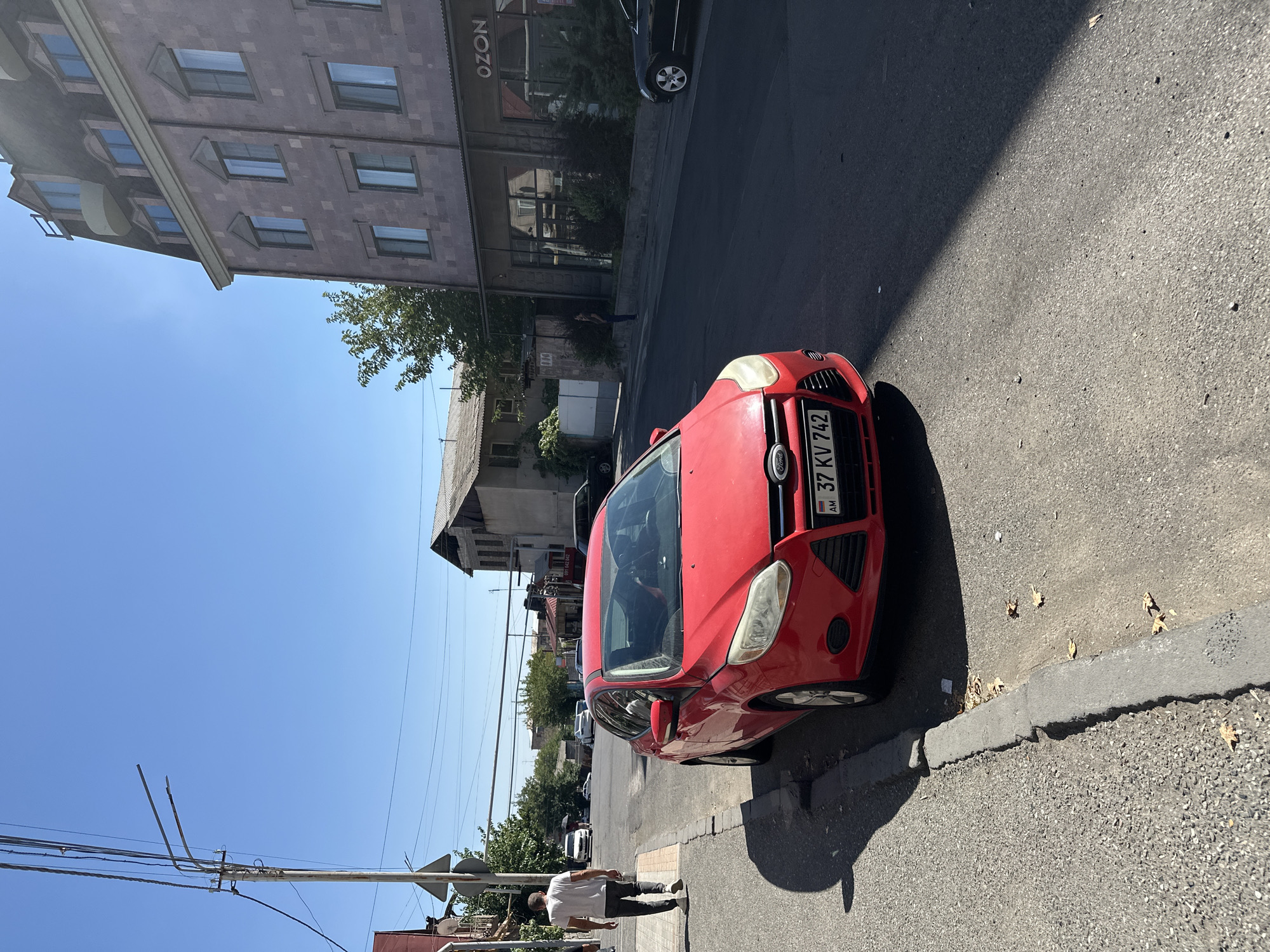 A red Ford Focus is parked on the side of a street in Yerevan, Armenia. The car, license plate AM 37 KV 742, faces right. A man in a white t-shirt and dark pants walks away from the camera on the left sidewalk. In the background, a multi-story building with arched windows stands on the right side of the street. The sky is clear and blue. The street is paved, and a concrete curb separates the road from the sidewalk. There are trees and other buildings visible in the distance.