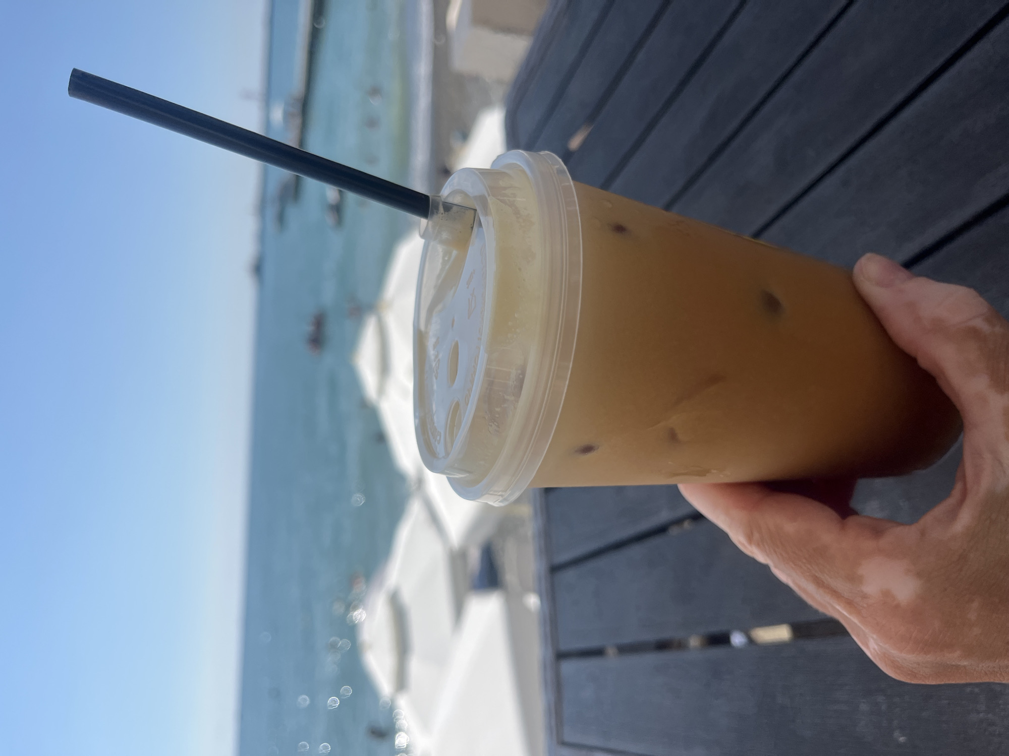 A hand with vitiligo holds a plastic cup with a lid and a black straw filled with iced coffee. The cup is placed on a dark wooden table. In the background, the turquoise sea meets a clear blue sky, with several white beach umbrellas and a pier visible. The scene suggests a sunny day at a beach resort, likely in Sochi, Russia, given the pier and typical resort architecture.