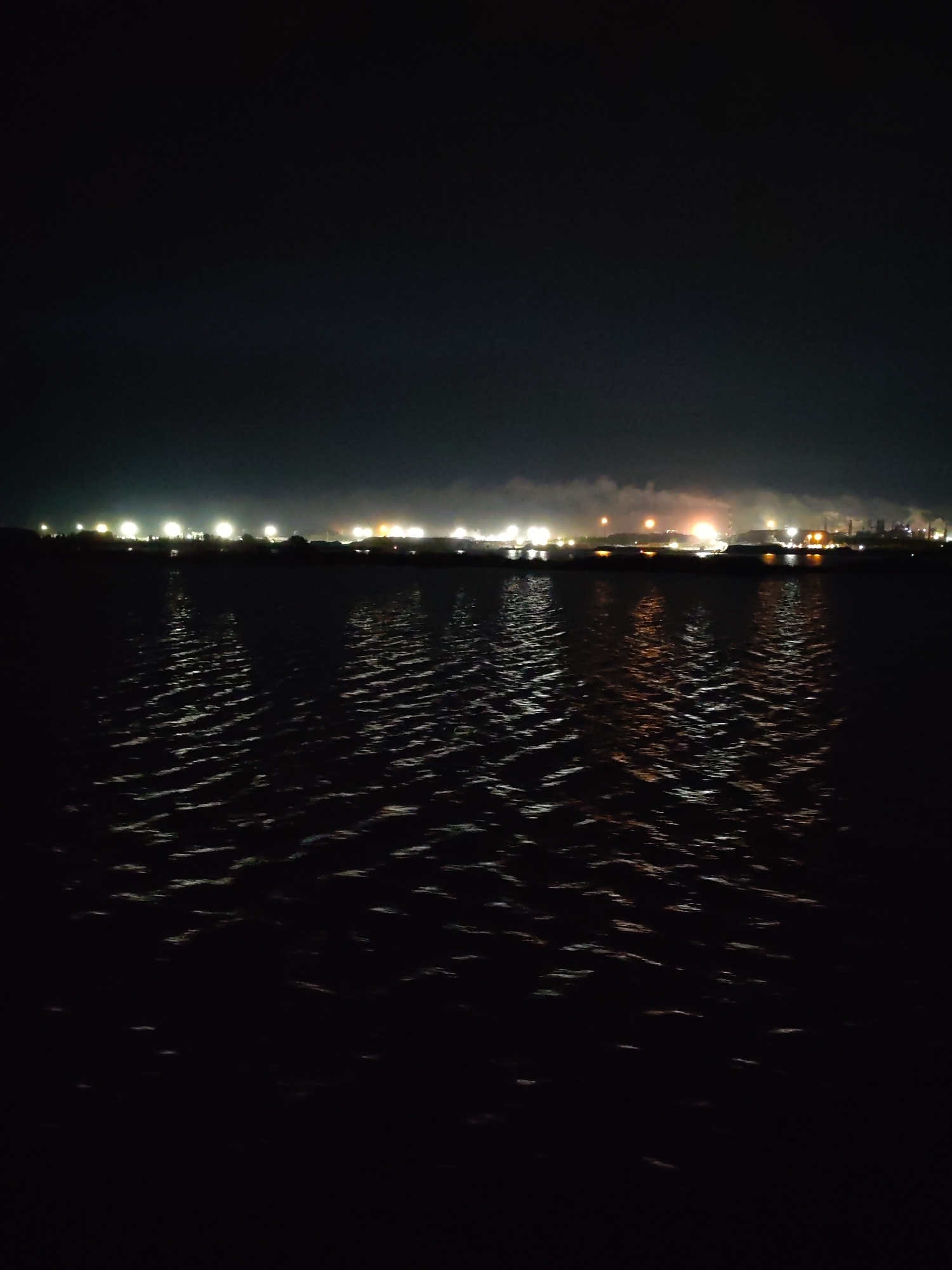 A night scene shows an expanse of water with subtle ripples, reflecting lights from a distant shoreline. The shoreline appears to be an industrial area with clustered lights and indistinct structures. Above the shoreline is a dark, featureless sky. No people are visible. The location is Unknown, Unknown.