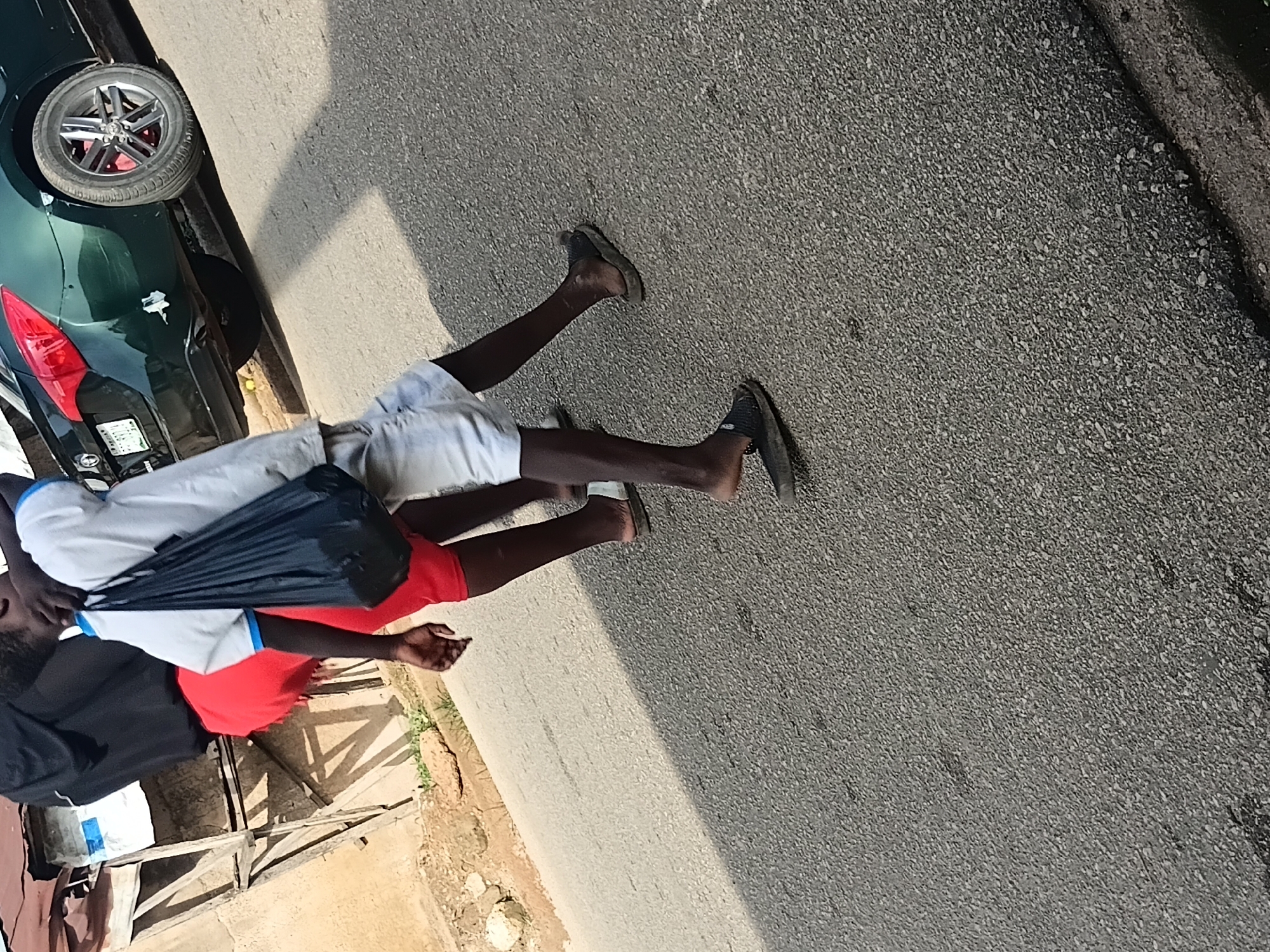 Two individuals are walking on a paved road. The person in the foreground is wearing white shorts and a white t-shirt, carrying a black plastic bag over their right shoulder. They are wearing dark sandals on their feet. Behind this person, another individual is visible wearing red shorts and a dark top. The background shows a green car parked on the side of the road, with a license plate that reads "SH 497DA". Part of a building and some wooden structures are also visible. The lighting suggests it is daytime, with shadows cast on the road.