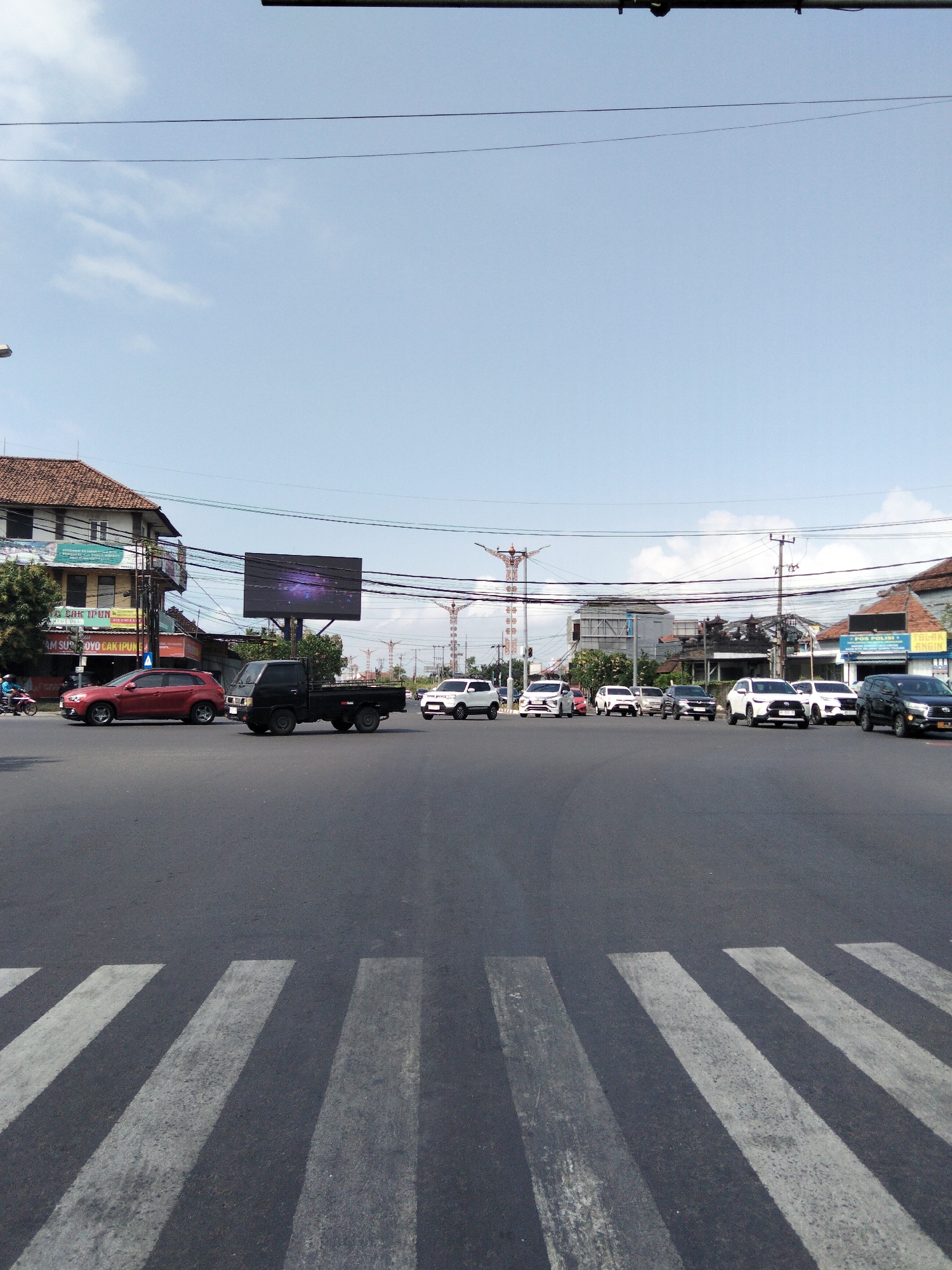 This is a full-frame, eye-level, natural light photograph of a street intersection in Denpasar, Indonesia, under a partly cloudy sky. In the foreground, a white-lined pedestrian crossing is visible on the asphalt road. Across the intersection, several vehicles are stopped or moving slowly. On the left, a red SUV is waiting, and to its right, a black pickup truck is stopped. Further in the distance, a line of white SUVs and other cars are present. Buildings line the sides of the street, including a multi-story building with a sign that appears to read "ATM SUYO CAK IPUN" and a large digital billboard displaying a purple galaxy image. Overhead, power lines crisscross the sky. The overall scene depicts a typical urban traffic situation in Denpasar.