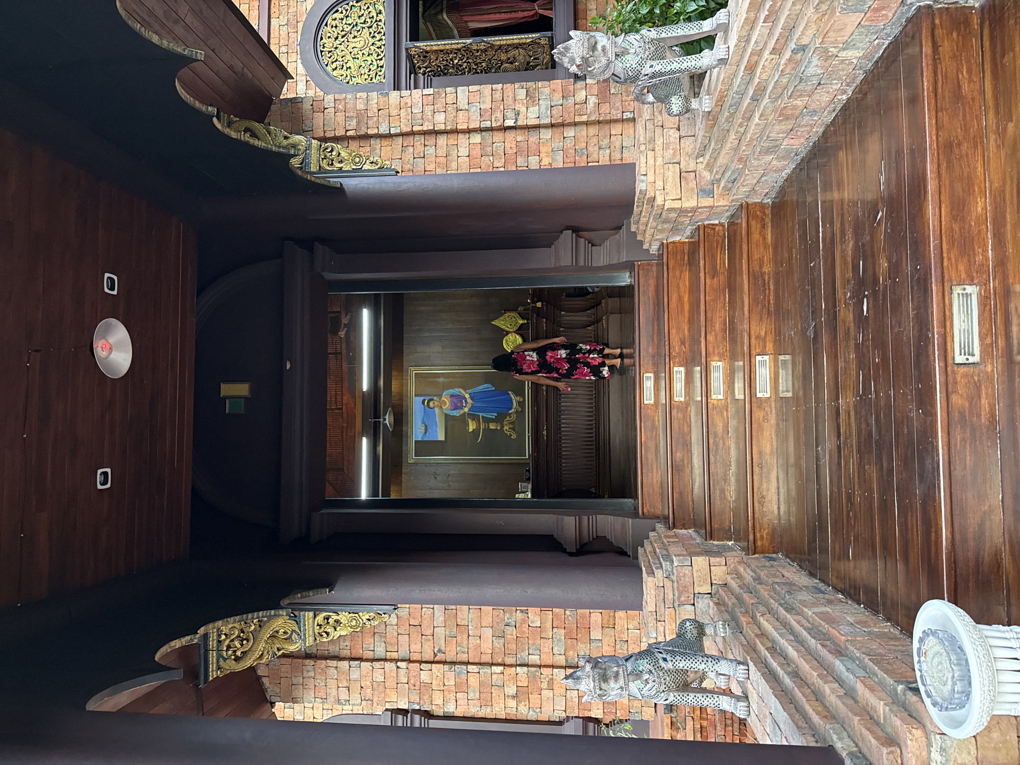 A person, seen from behind and wearing a floral dress, walks up a wide wooden staircase towards the entrance of a building. The entrance is framed by dark wooden columns and brickwork, with ornate golden carvings on the archways. Flanking the staircase are two stone lion-like statues. Inside the building, a painting of a woman in traditional attire is visible, along with some decorative items. The building appears to be a traditional Thai architectural style, possibly a temple or a historic residence, located in the Ko Samui District of Thailand. The ground is paved with wooden planks, and the walls are constructed with red bricks. An ashtray sits on a white pedestal in the foreground.