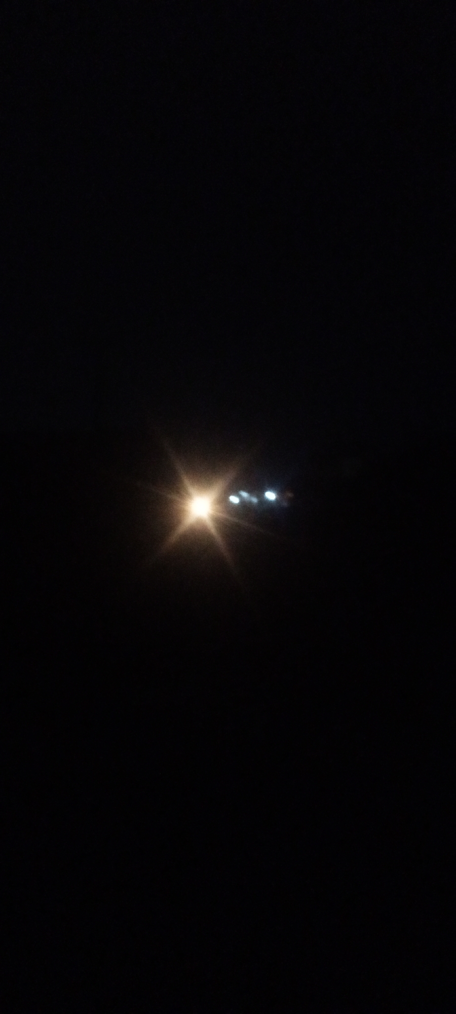 A nighttime scene showing light sources in the darkness. The scene is likely at the city of Sabon Tasha, Nigeria. The light sources include a bright starburst of light with smaller lights nearby. The surrounding environment is dark and indistinct.