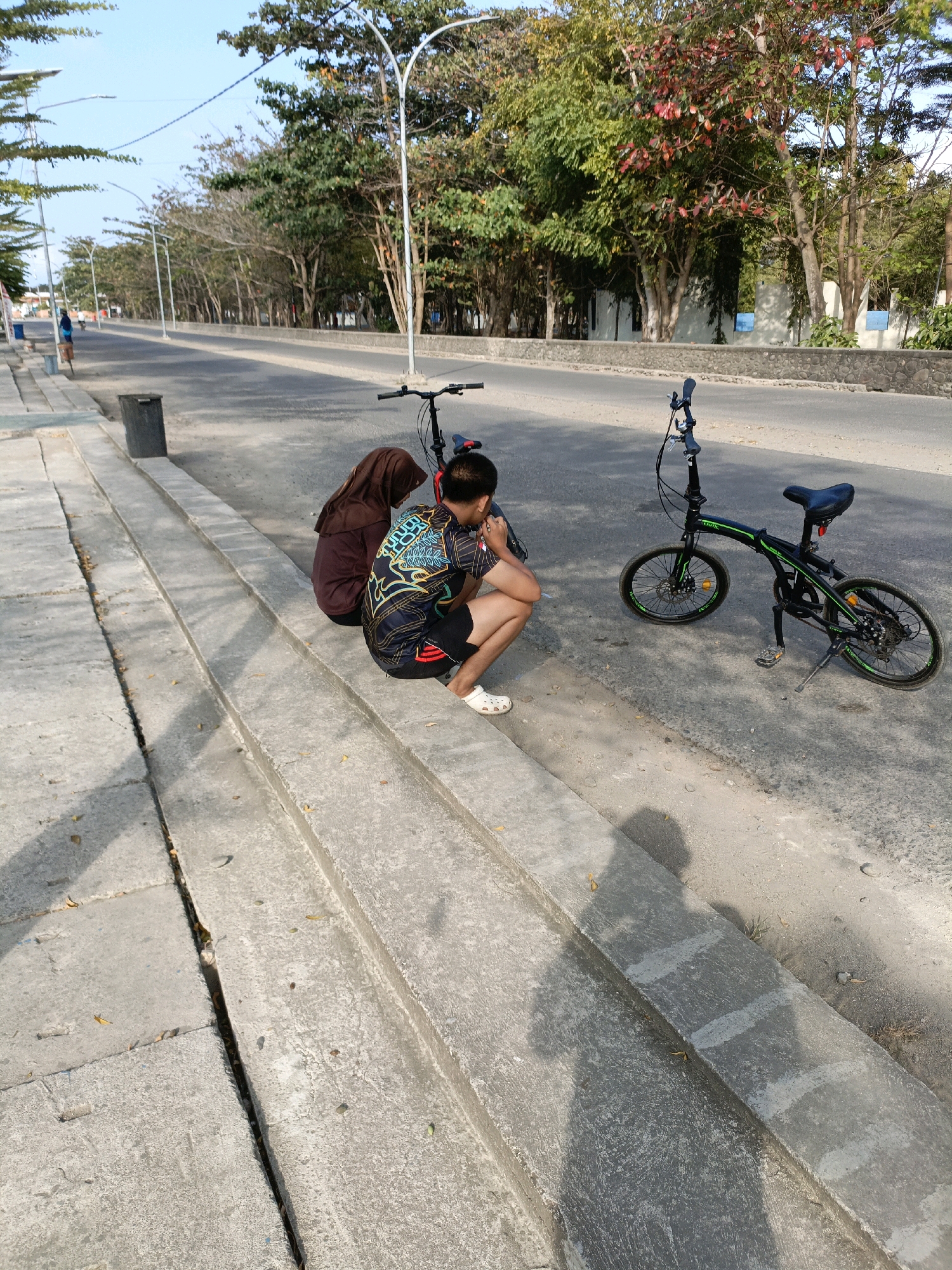 The image captures a sunny day on a paved road in Bulukumba, Indonesia, likely a coastal area given the presence of palm-like trees and a clear sky. In the foreground, two individuals are seated on concrete steps adjacent to the road. One person, wearing a brown hijab and a dark garment, is facing away from the camera. Beside them, another individual, dressed in a black and colorful patterned t-shirt and black shorts, is crouching and looking down. Two foldable bicycles are parked on the side of the road. One is closer to the seated individuals, and the other is further down the road. The environment includes a sidewalk, a trash can, streetlights, and a line of trees along the roadside. The overall scene suggests a leisurely break during an outdoor activity.
