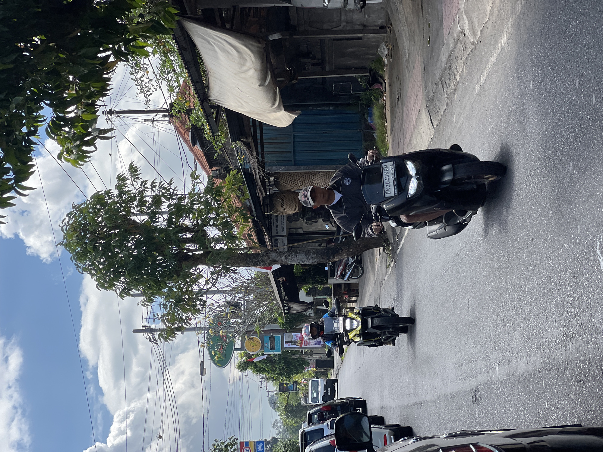 The image depicts a street scene in Ubud District, Indonesia, on a bright, partially cloudy day. The road is paved and appears to be a moderately busy thoroughfare with several vehicles. In the foreground, two individuals are riding motorcycles. The motorcycle on the right is a dark-colored scooter, and the rider is wearing a dark jacket and a baseball cap. The motorcycle on the left is a yellow adventure-style bike, and the rider is wearing a blue jacket and a helmet. Both motorcycles have license plates visible.
To the right of the road, there are buildings with corrugated metal roofs and weathered facades, some with awnings or tarps. Vegetation, including trees with green foliage, is present throughout the scene, with branches extending over the road and sidewalks. Utility poles with numerous electrical wires are visible against the sky. Signs for businesses are also present along the street. The overall environment suggests a typical street scene in a tropical urban area, likely a commercial or mixed-use district within Ubud.