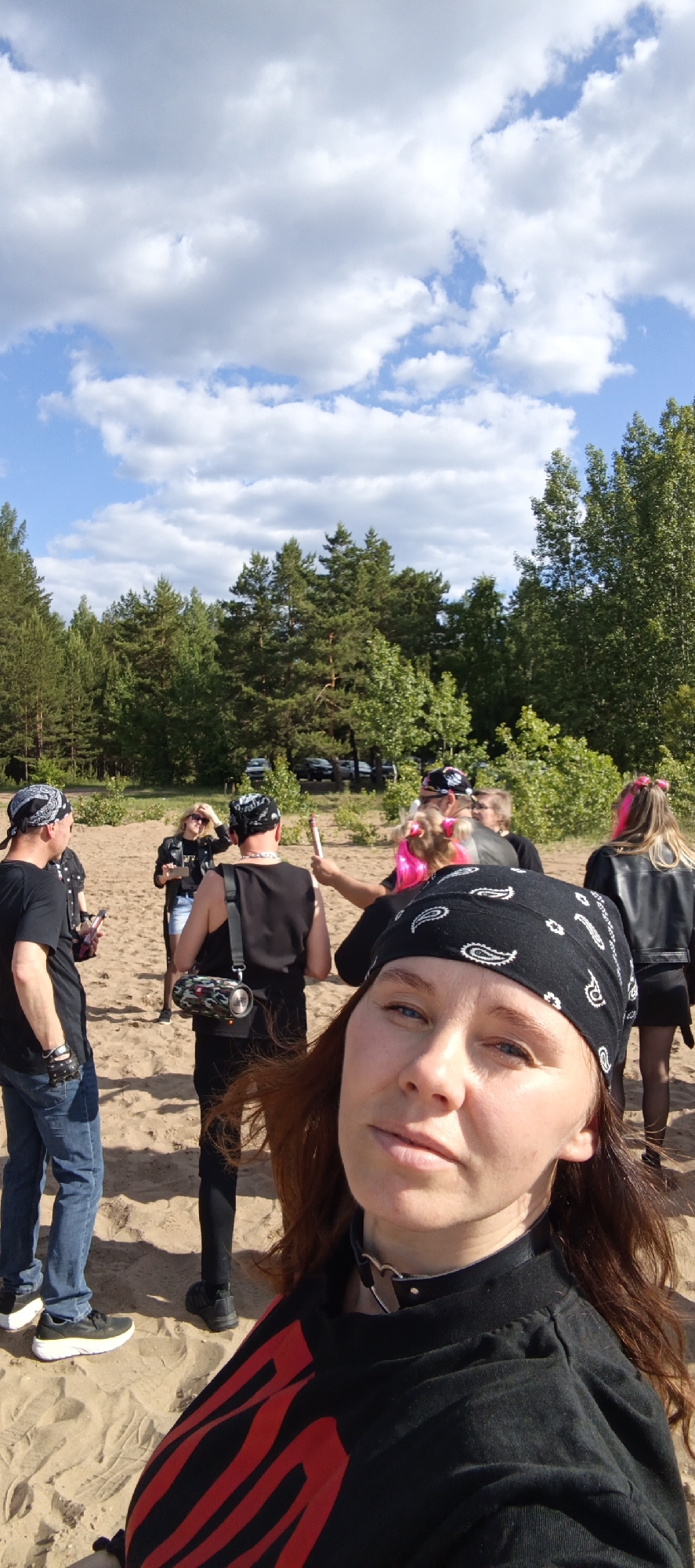 An outdoor scene in Noviy, Russia, depicts a group of individuals gathered on sandy terrain under a partly cloudy blue sky. In the immediate foreground, a woman with long reddish-brown hair captures a selfie, looking directly at the camera. She wears a black bandana with white paisley patterns, a black t-shirt featuring red diagonal patterns on the chest, and a black choker with a metallic heart-shaped buckle.
Seven additional individuals are visible in the midground, positioned on the sand. The majority are dressed in black clothing and bandanas. To the far left, a man in a black t-shirt, blue jeans, and black fingerless gloves holds a mobile device. Centrally, a man in a black tank top and bandana carries a camouflage-patterned JBL portable speaker slung over his shoulder; his back is to the camera. Adjacent to him, another individual, also facing away, exhibits vibrant pink hair extensions and holds a long, cylindrical object with a red tip. To the right, a woman is dressed in a black leather jacket and a black skirt, with long blonde hair featuring pink highlights.
The background consists of a dense forest line comprising coniferous and deciduous trees. Beyond the trees, several dark-colored vehicles are parked, indicating an adjacent road or parking area. The sandy ground is largely barren, with minimal scattered low-lying vegetation.