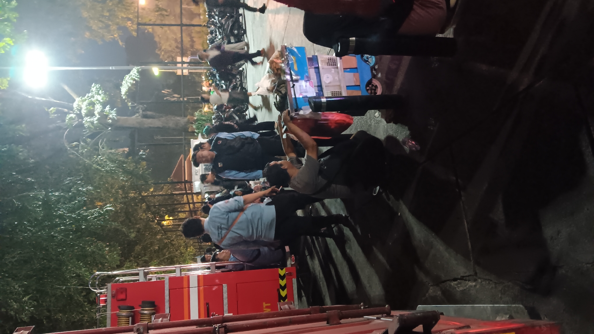 This is a medium shot, taken at night, of a group of people gathered in what appears to be an outdoor parking area or street in Central Jakarta, Indonesia. The scene is illuminated by artificial lighting from street lamps and lights on a red emergency vehicle, possibly a fire truck, visible on the left side of the frame.
In the foreground, a man wearing a grey t-shirt and dark pants is crouching down next to a small, blue and white wheeled cart laden with items, some of which are in plastic bags. He is holding a red plastic bag in his right hand. The ground is paved with cracked asphalt, and long shadows are cast by the people and objects.
Behind the crouching man, several other individuals are standing and interacting. One man in a light blue shirt with an orange strap across his chest is facing away from the camera. Another man in a dark vest over a blue shirt and a dark cap is looking towards the right. Several other people are visible in the middle ground, some appearing to be in uniform or work attire.
In the background, a large number of motorcycles are parked, suggesting this is a public area or event. Trees with dense foliage are present, and a light pole with a bright lamp stands tall in the background, contributing to the nighttime ambiance. The overall impression is one of a gathering or event taking place after dark, with the emergency vehicle possibly indicating a service or response.