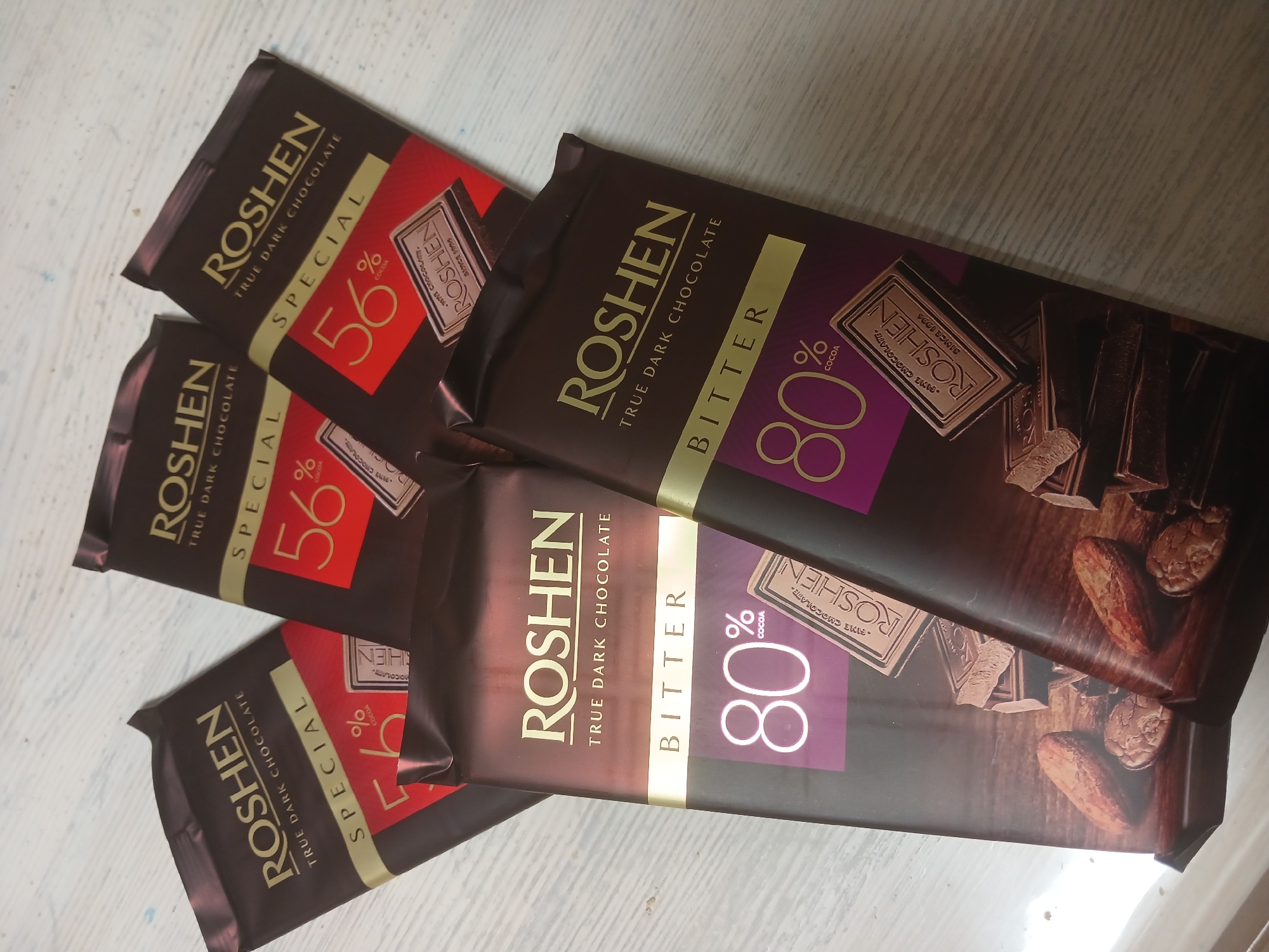 The image shows a top-down view of several bars of Roshen chocolate on a light-colored wooden surface. Four of the bars are partially visible, with the focus on two prominent bars in the foreground. These foreground bars are labeled "ROSEN TRUE DARK CHOCOLATE BITTER 80% COCOA" and display an image of broken chocolate pieces and cocoa beans. Two other bars in the background are labeled "ROSEN SPECIAL 56% COCOA". The scene is indoors, with no indication of a specific location in Sumy, Ukraine.