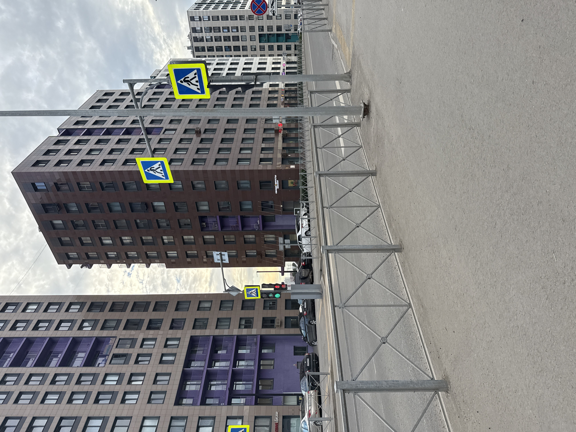 The image depicts an urban street scene in Perm, Russia, with modern high-rise apartment buildings lining the road. Two prominent pedestrian crossing signs, each a blue square with a white pictogram of a person walking, are visible, one mounted on a pole extending over the sidewalk and another on a separate pole. A traffic light at the intersection shows a green signal. Several cars are parked along the street and on the side of the road. A metal barrier with a diamond pattern separates the sidewalk from the road. The sky is overcast, suggesting cloudy weather. The buildings feature a mix of brown and beige facades with purple accents on balconies and window frames. The overall impression is one of a contemporary residential area in a Russian city.