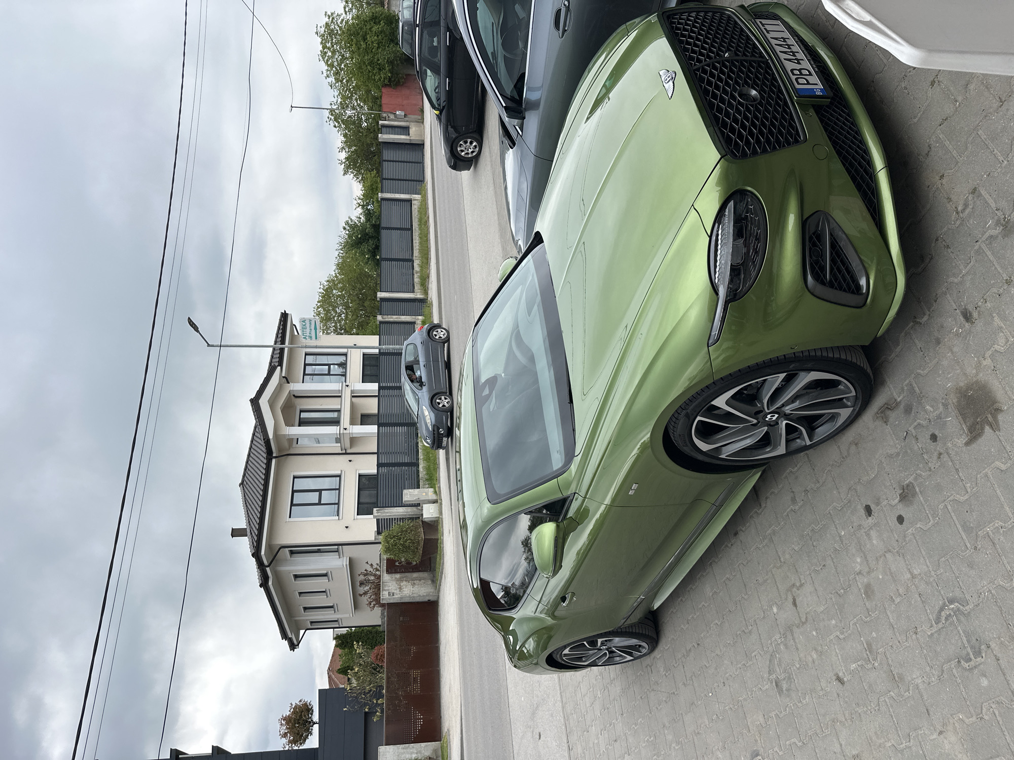 A green Bentley Continental GT is parked on a paved street in a residential area of Plovdiv, Bulgaria. The license plate number PB 4444 TT is visible. A modern, two-story house is on the left side of the frame. Two other cars, one gray and one black, are parked behind the Bentley. Utility poles and overcast skies are present. No people are clearly visible in the immediate vicinity of the car.