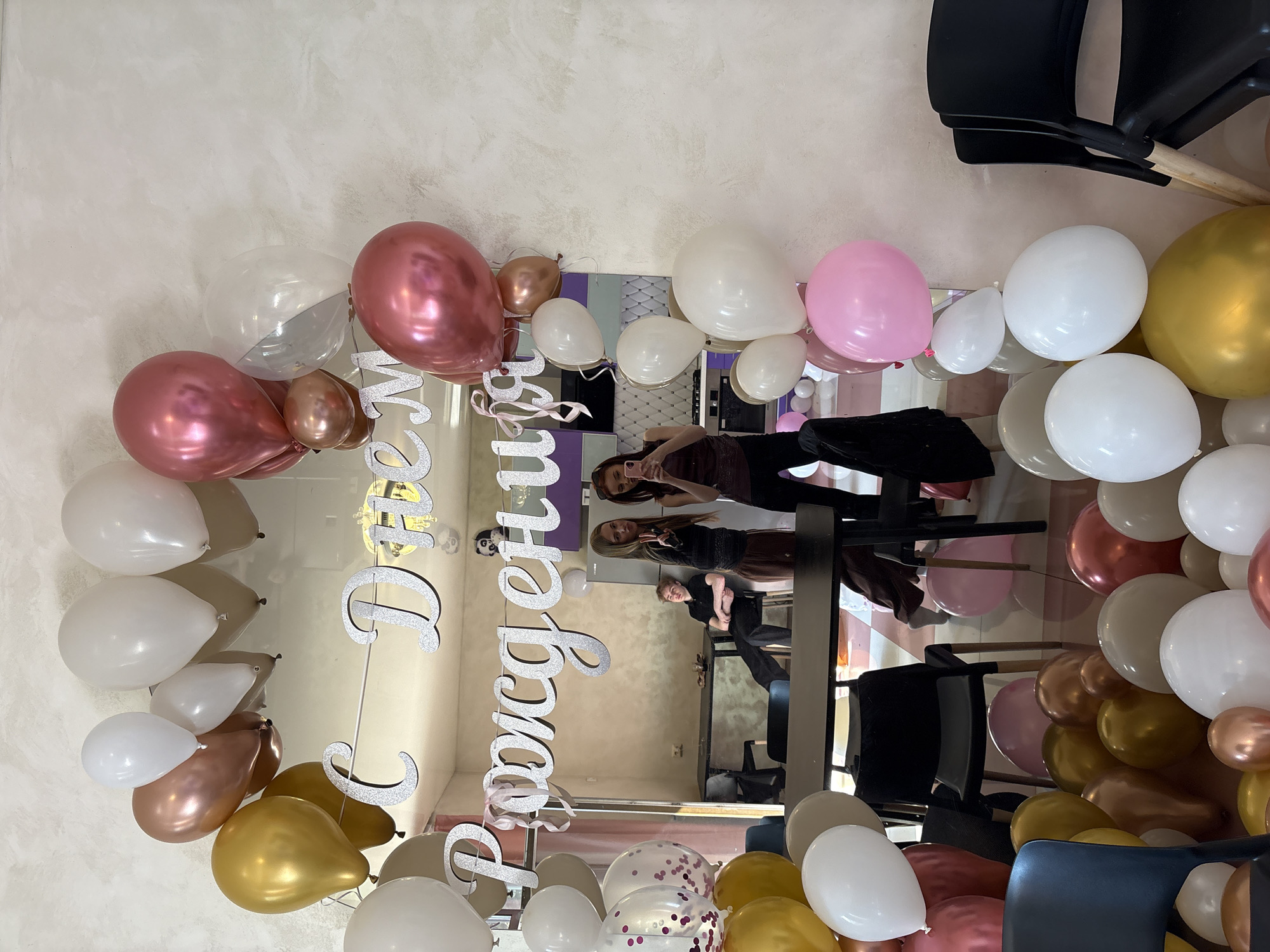 The image shows a reflection in a mirror, surrounded by balloons of various colors (white, pink, gold, and rose gold) and a banner in Russian that reads "Happy Birthday". Three women are reflected in the mirror. The environment appears to be indoors, possibly a salon or event space. Based on the banner language, the location is likely in Russia. I am unable to confirm that the image is located specifically in Saint Petersburg, Russia.