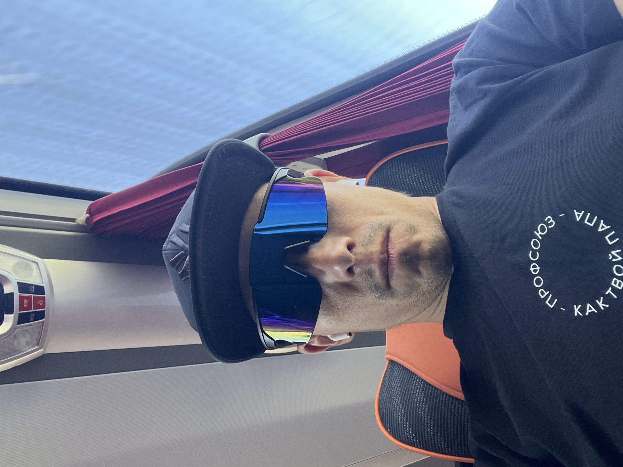 A man wearing a black baseball cap and blue reflective sunglasses is seated inside a vehicle. The vehicle appears to be a bus or train, indicated by the overhead lighting and passenger control panel on the left. Red curtains are visible above the window. The man is wearing a black shirt with Cyrillic text in a circular pattern. The location is Chaikovsky, Russia, based on the Cyrillic text.