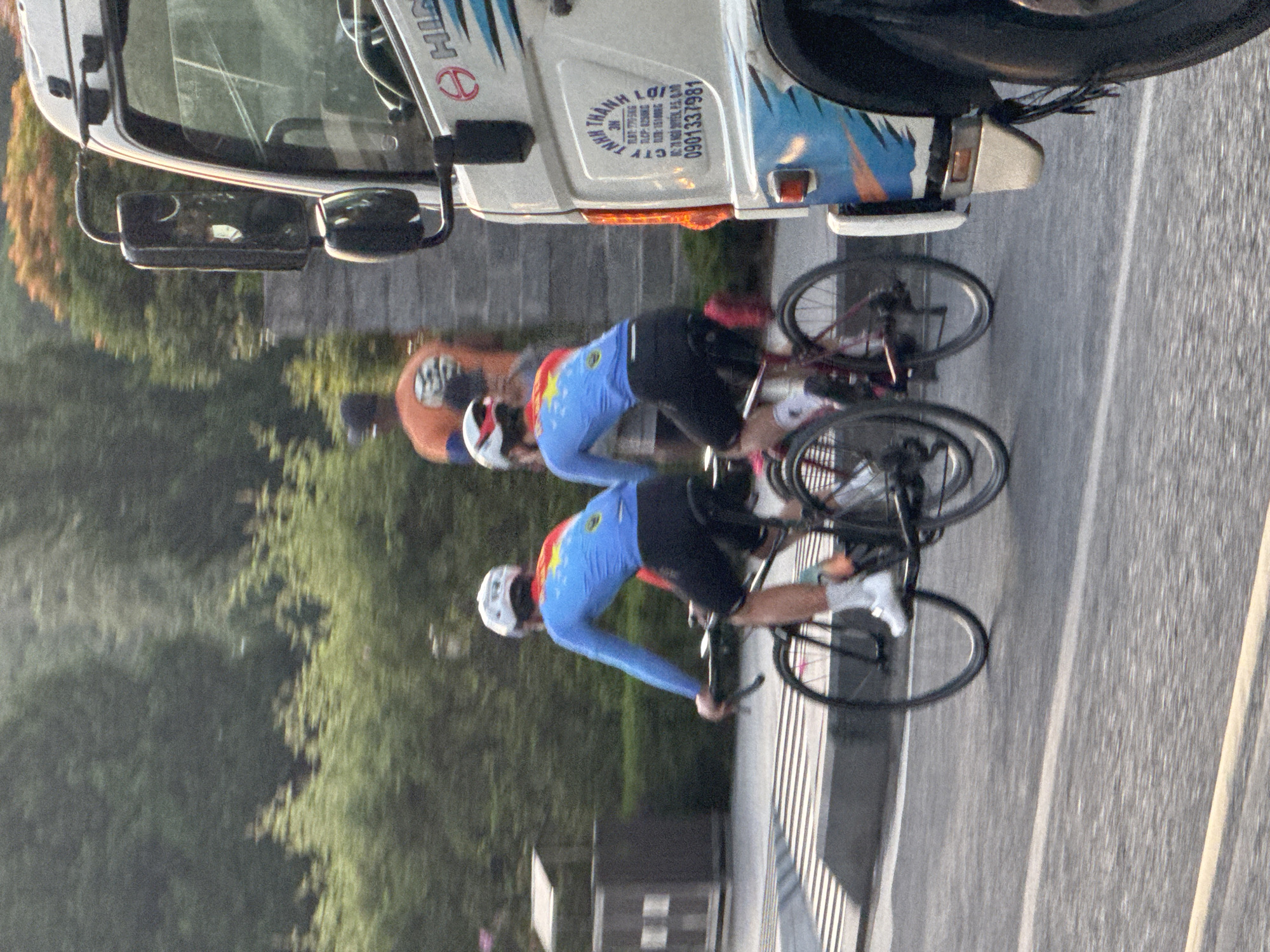The image depicts a street scene in Da Lat, Vietnam, featuring two cyclists in bright blue and red cycling jerseys riding on a road. They are wearing white helmets and black cycling shorts, with white socks and shoes. The cyclist on the left has a white helmet, while the cyclist on the right has a white helmet with red accents. Behind them, another person in an orange shirt and a person in a dark shirt are visible, slightly out of focus. A white truck with blue and orange graphics is prominently positioned on the right side of the frame, partially obscuring the cyclists and the background. The truck has visible branding, including the HINO logo and Vietnamese text that appears to be a license plate or company information. The environment suggests a slightly overcast day, with trees and foliage visible in the background. The road surface is asphalt, with distinct white lines and a textured crossing area in the foreground. The overall impression is of a moment captured during a cycling event or activity on a city street.