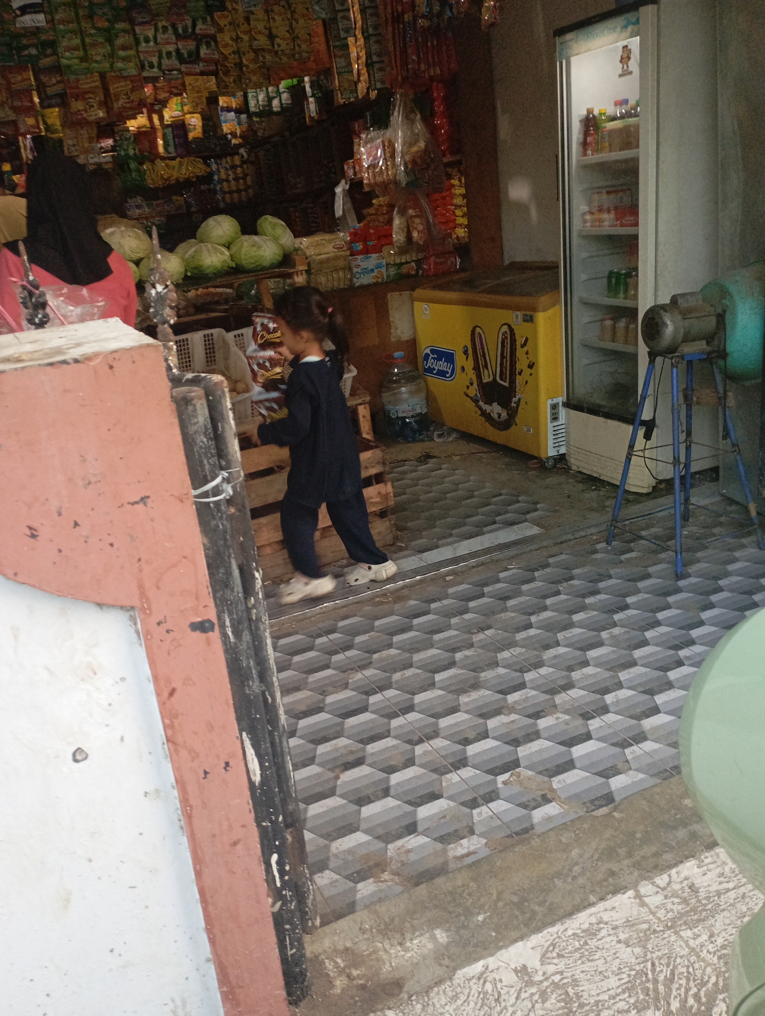 In Kecamatan Haurgeulis, Indonesia, an indoor scene depicts a small convenience store. A young girl, wearing a dark blue long-sleeved shirt and pants, stands in the foreground to the right, walking towards the left. She is wearing light-colored sandals. To her left is a large, salmon-colored structure with dark vertical elements. Behind the girl, wooden crates are visible, and on top of them, a variety of products, including what appear to be cabbages and packaged goods, are displayed. Further to the right, a white upright refrigerator is open, revealing shelves stocked with bottles and containers. In front of the refrigerator is a yellow chest freezer with a cartoonish design of ice cream cones. A blue metal stand with a motor-like object is positioned near the freezer. The floor is covered with patterned hexagonal tiles. The background is filled with shelves stocked with numerous packaged food items and snacks. The overall impression is of a well-stocked, local grocery store.