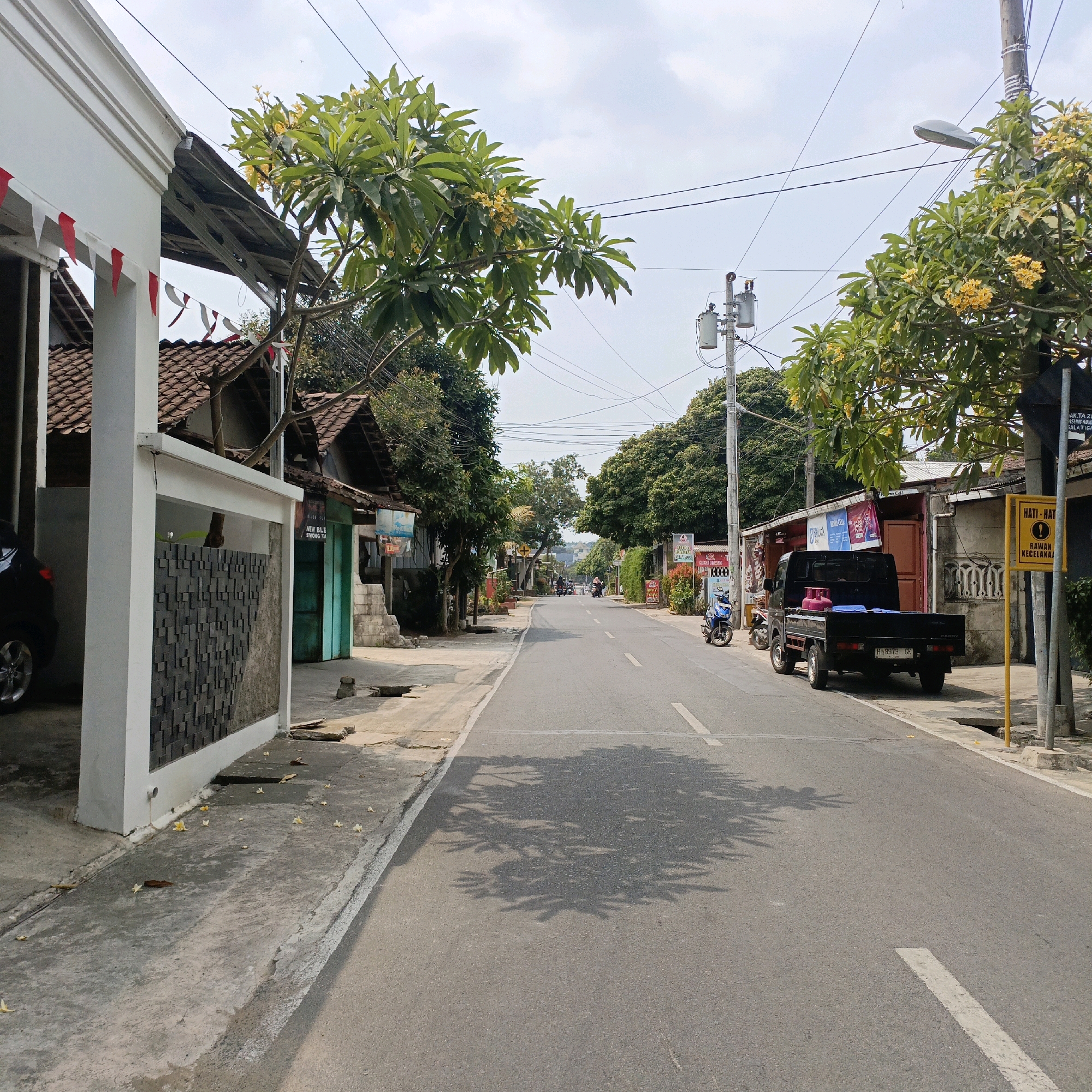 A street scene in Salatiga, Indonesia, shows a paved road lined with buildings and trees. A black pickup truck with a pink gas tank in the bed is parked on the right side of the road. A blue motorcycle is parked behind it. A yellow sign with red text reading "HATI-HATI" ("Beware") and an exclamation mark inside a yellow triangle is visible on a pole on the right side of the road. The street is clear of heavy traffic, with a few motorbikes and people visible in the distance. The buildings appear to be a mix of shops and residences, some with tiled roofs. Overhead power lines crisscross the sky. The overall scene depicts a typical street in a bustling Indonesian city.