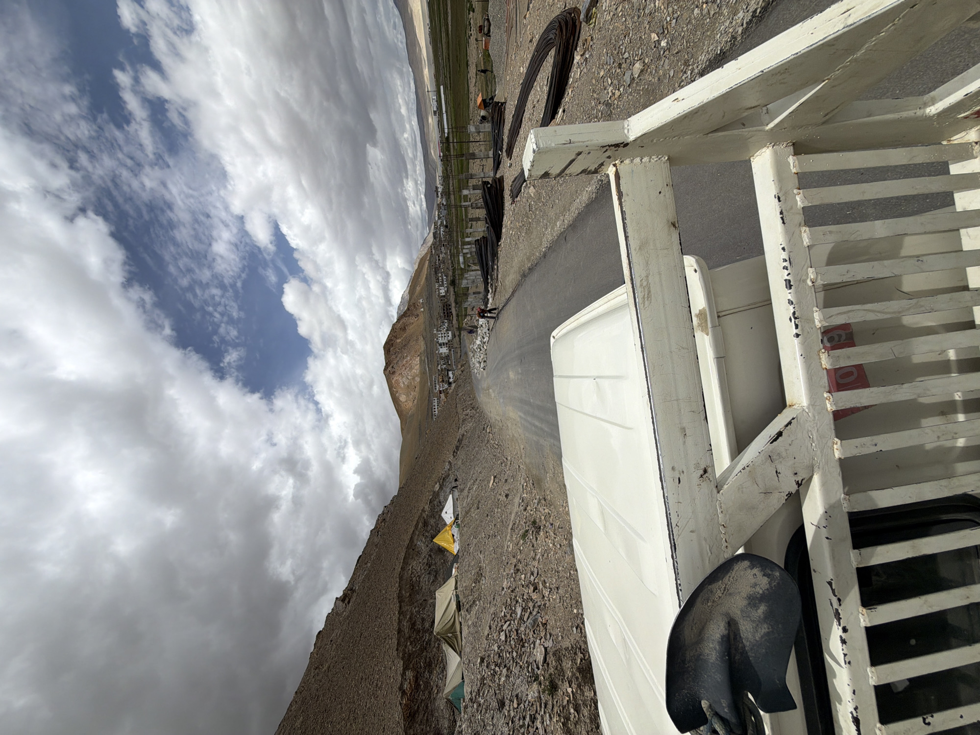 The image shows a view from the back of a white truck, looking forward along a paved road in Karzok, India. On the left side of the road, several tents are set up on a rocky, sloped terrain, with a prominent yellow tent. Further up the road, buildings of a settlement are visible. On the right side of the road, there is a construction site with piles of rebar and some concrete pillars. A few individuals are present at the construction site. The sky is partly cloudy with patches of blue. The overall environment suggests a remote, high-altitude location.