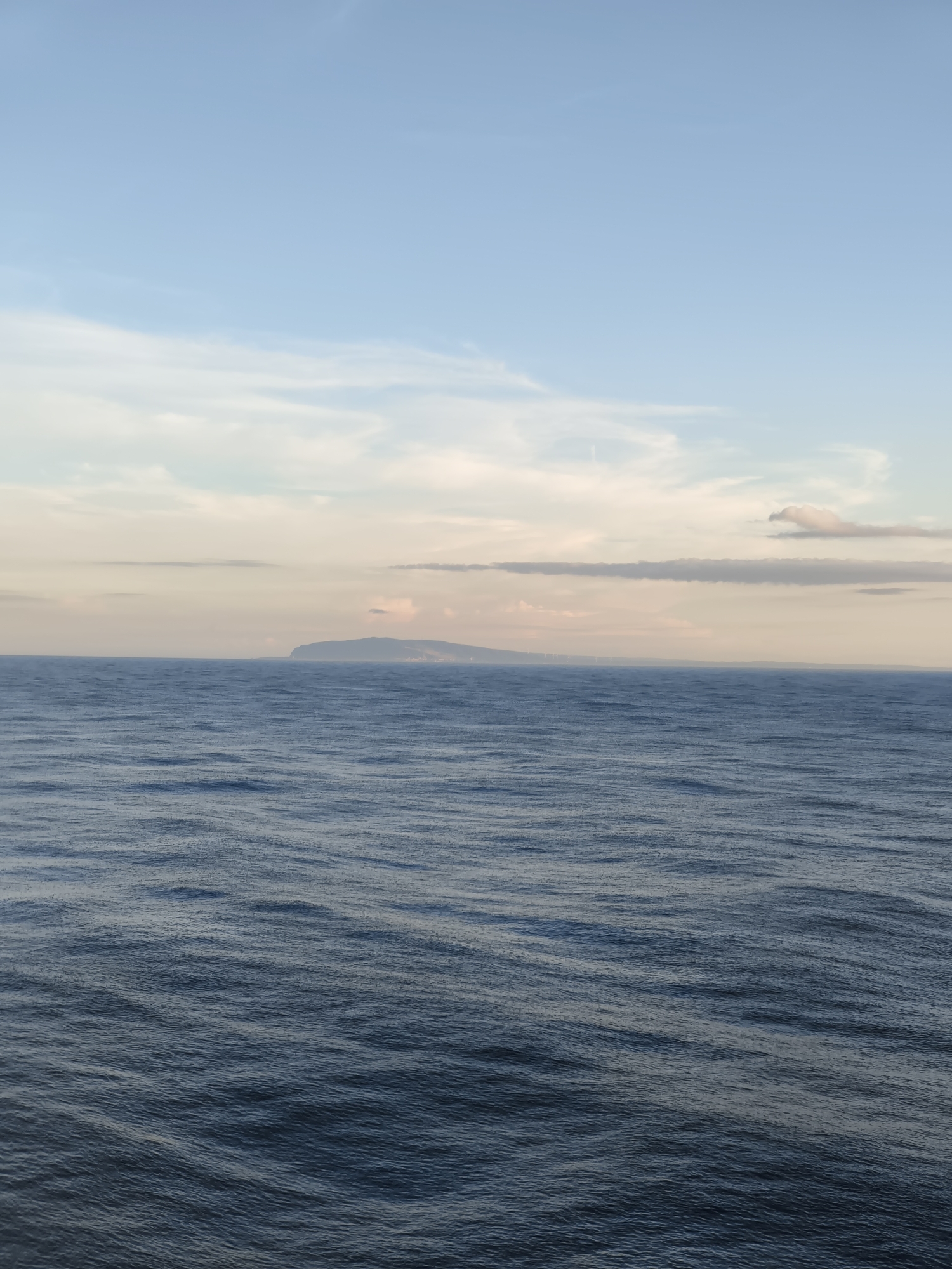 A vast expanse of deep blue ocean stretches across the lower half of the frame, its surface rippled with gentle waves that catch the light. In the distance, a hazy, low-lying island or coastline is visible on the horizon, its shape softened by the atmospheric perspective. Above the ocean, the sky transitions from a pale, almost ethereal light near the horizon, where wisps of pink and yellow clouds are present, to a clear, soft blue at the top of the image. The overall impression is one of calm and open space, with the distant landmass providing a subtle focal point.