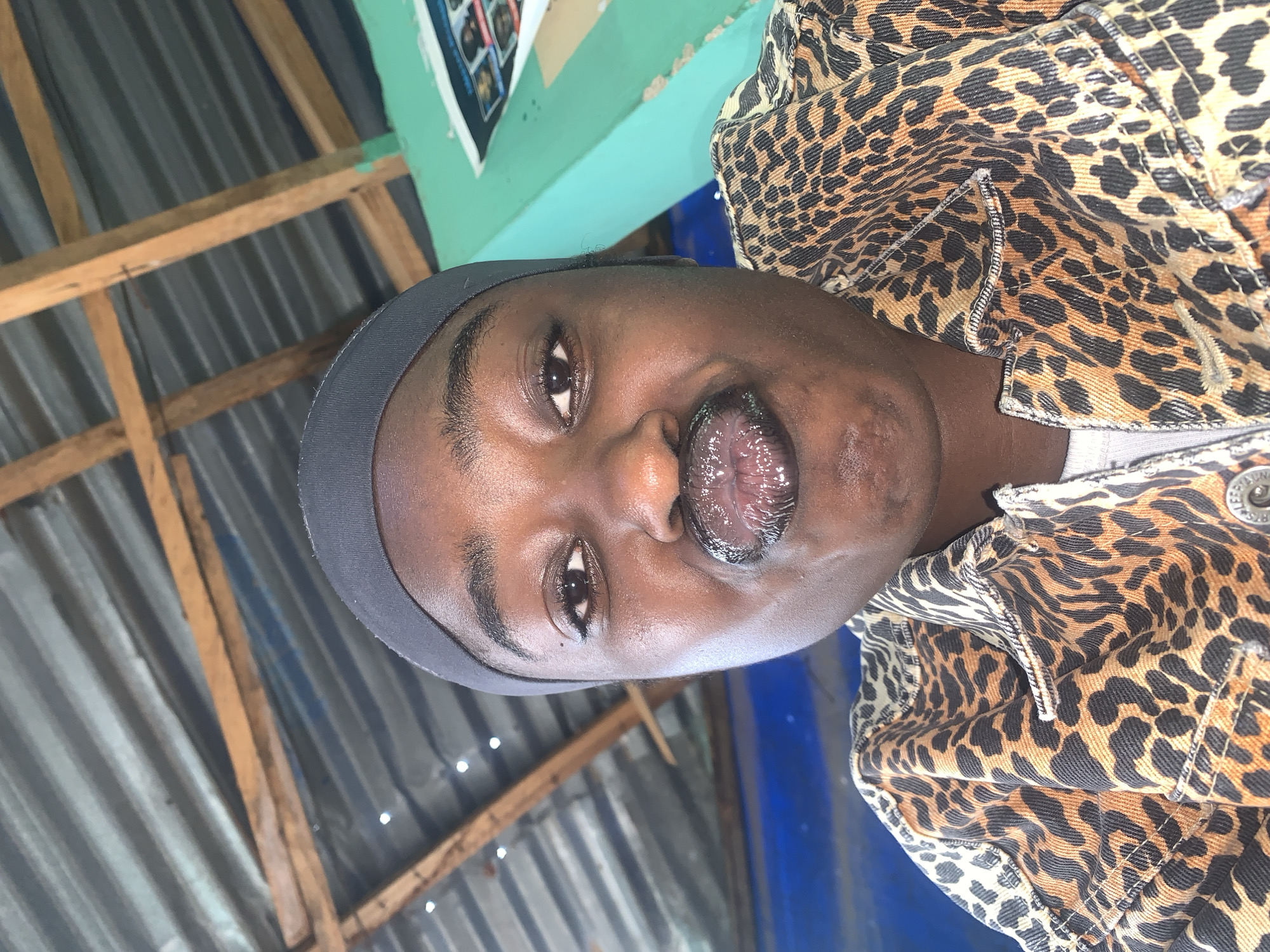 A person with dark skin is wearing a black headband and a leopard-print jacket. The person's tongue is extended and has a glossy appearance. The background includes a corrugated metal roof and wooden beams. A turquoise wall with an object attached to it is also visible in the upper portion of the image. The location is New Stadium, Nigeria.