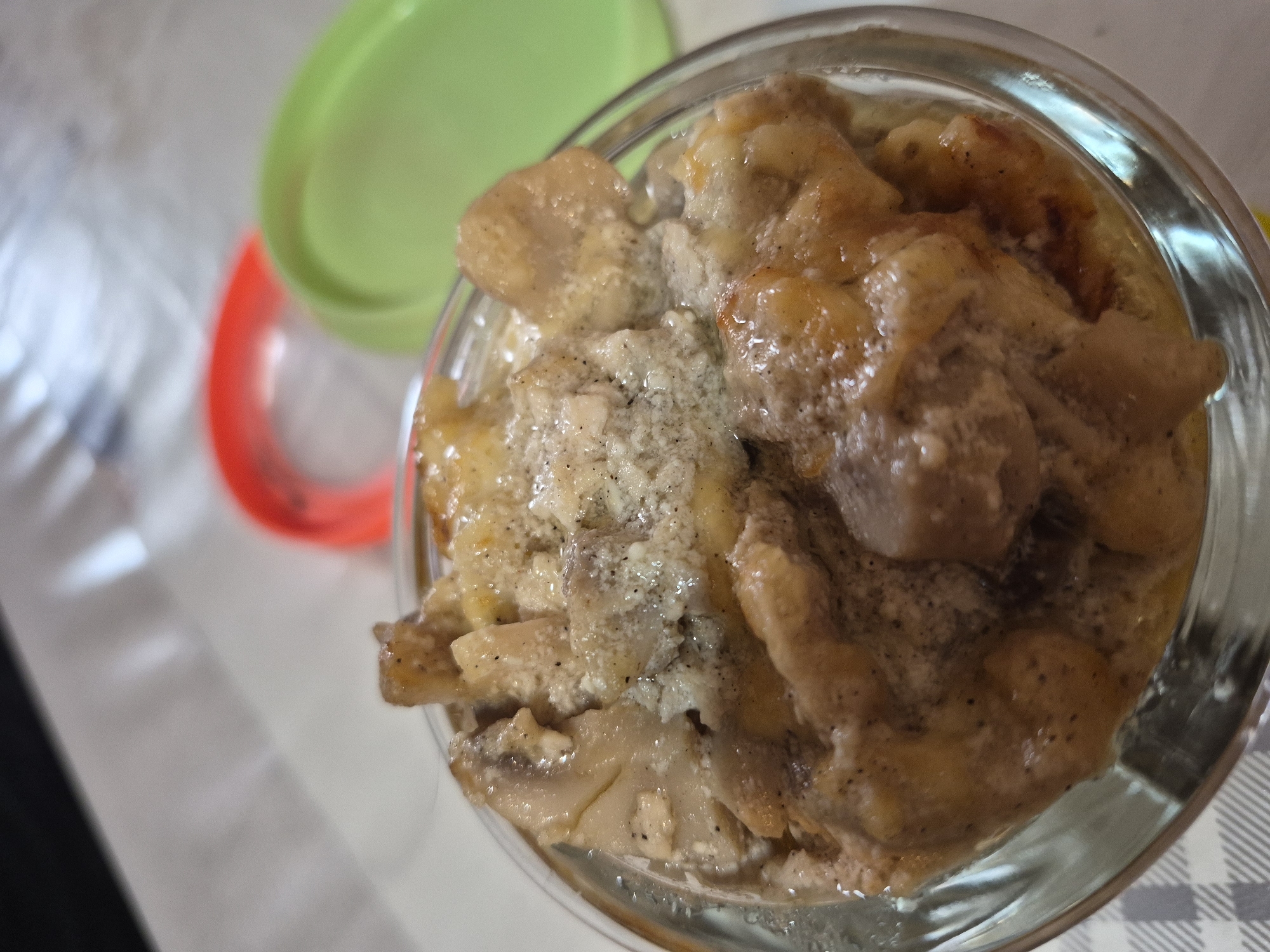 This is a close-up, top-down shot of a serving of food, possibly a casserole or gratin, in a clear glass dish. The dish is filled with a creamy, beige sauce with visible pieces of what appear to be mushrooms and possibly chicken or other white meat. The food is seasoned with black pepper. In the background, out of focus, are two plastic lids, one red and one green, suggesting this might be a prepared meal in a take-out container. The setting appears to be indoors, with a light-colored, textured surface visible beneath the dish, possibly a tablecloth or placemat with a checkered pattern. The lighting suggests it is daytime. There is no identifiable location within the city of Yerevan, Armenia.