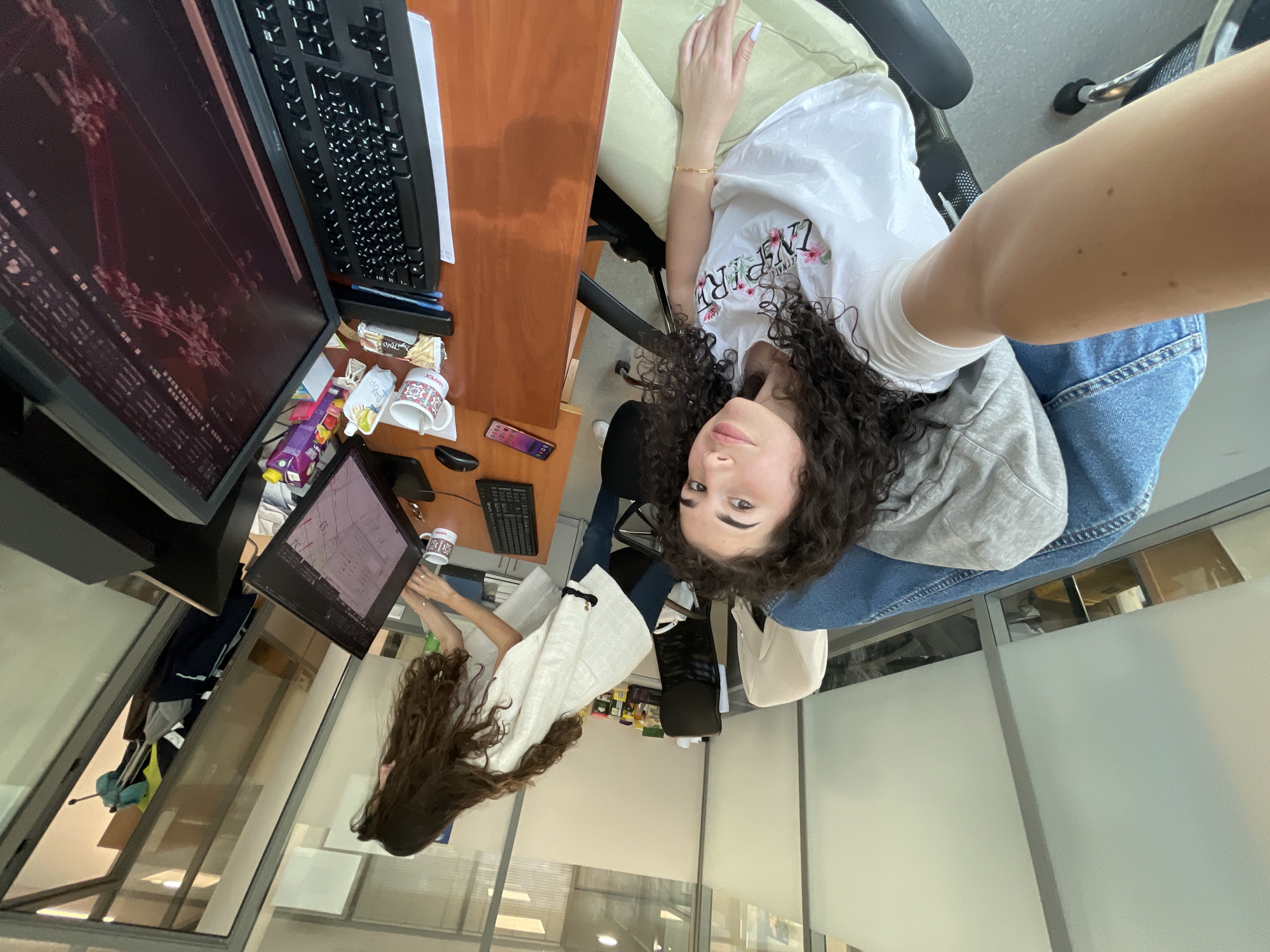 A selfie-style photograph taken indoors at an office or workspace. The presumed photographer is a young woman with curly dark hair. She's wearing a white t-shirt with a floral design and is seated in a chair. Her arm extends into the frame. Part of her denim jeans and a gray hoodie are visible. In the background and to the left, there is a desk. On the desk, there is a monitor displaying what appears to be technical drawings or schematics. To the right of the monitor, there is a keyboard, a coffee mug, and miscellaneous office supplies, including snacks and a mobile phone. There is a second screen on a smaller stand and a second woman looking at it. The room has large windows that provide ample natural light, with frosted glass partitions that obscure the view to some extent.
It's impossible to verify the exact location as Sofia, Bulgaria without external information.