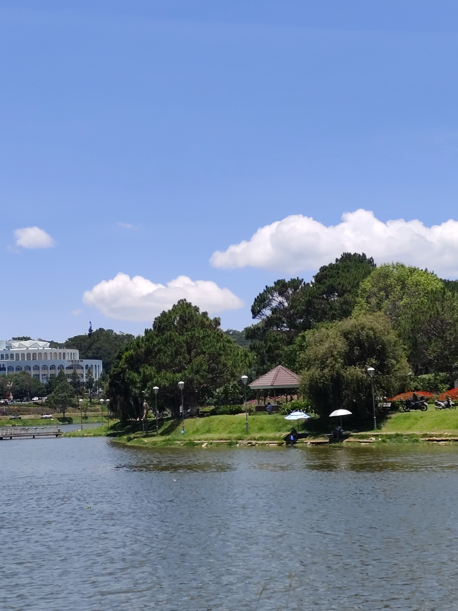 A vibrant daytime scene captures a panoramic view of a lake in Da Lat, Vietnam, under a clear blue sky dotted with fluffy white clouds. The calm, rippling water occupies the foreground and mid-ground of the image.
On the opposite shore, a lush green bank is visible, adorned with numerous trees, including tall pines and other broadleaf species. On the left side of the bank, a large, light-colored building with multiple arched windows and a distinctive roofline stands out, resembling a public building or resort. A small bridge or walkway extends into the water on this side.
Towards the center and right of the distant bank, two individuals are seated near the water's edge, seeking shade under a white umbrella and a blue-and-white patterned umbrella, appearing to be fishing or relaxing. Behind them, nestled among the trees, is a small gazebo with a brown roof. Several lampposts are spaced along the bank, and two motorcycles are parked on the grassy area on the far right. The bright sunlight and absence of shadows indicate a midday or early afternoon setting, conveying a serene and pleasant atmosphere. No discernible text is visible.