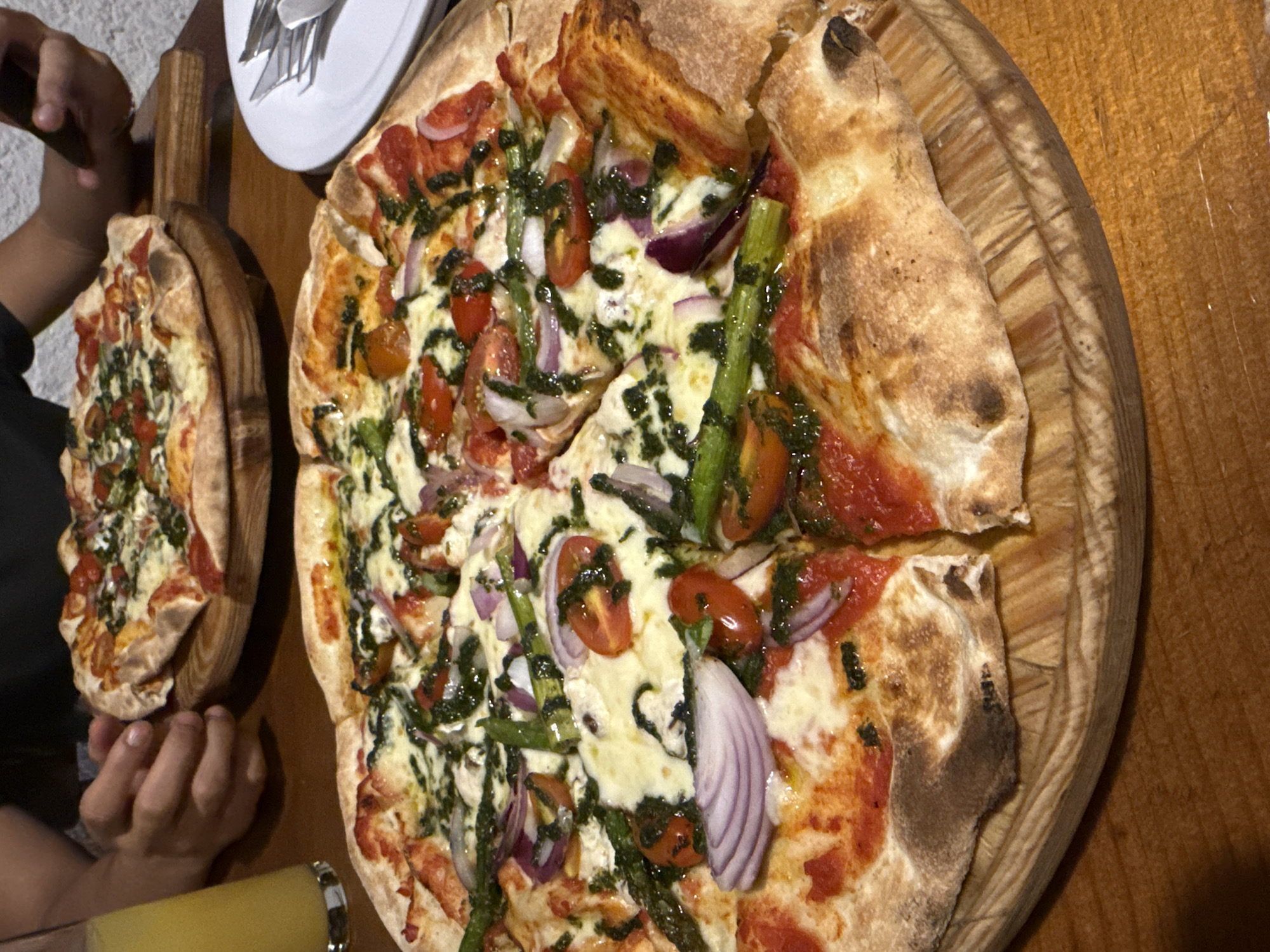 The image depicts two pizzas on wooden serving platters. The larger pizza, cut into slices, sits on a round wooden board, displaying toppings of tomato sauce, mozzarella, sliced tomatoes, red onion, asparagus, and a green drizzle, possibly pesto. The smaller pizza, also on a wooden platter, is topped similarly but appears to be smaller. Two hands are visible near the smaller pizza. A plate with silverware and a glass are present in the background. The setting appears to be indoors, likely a restaurant or dining area in Chipilo de Francisco Javier Mina (Chipilo), Mexico. The surfaces are wooden.