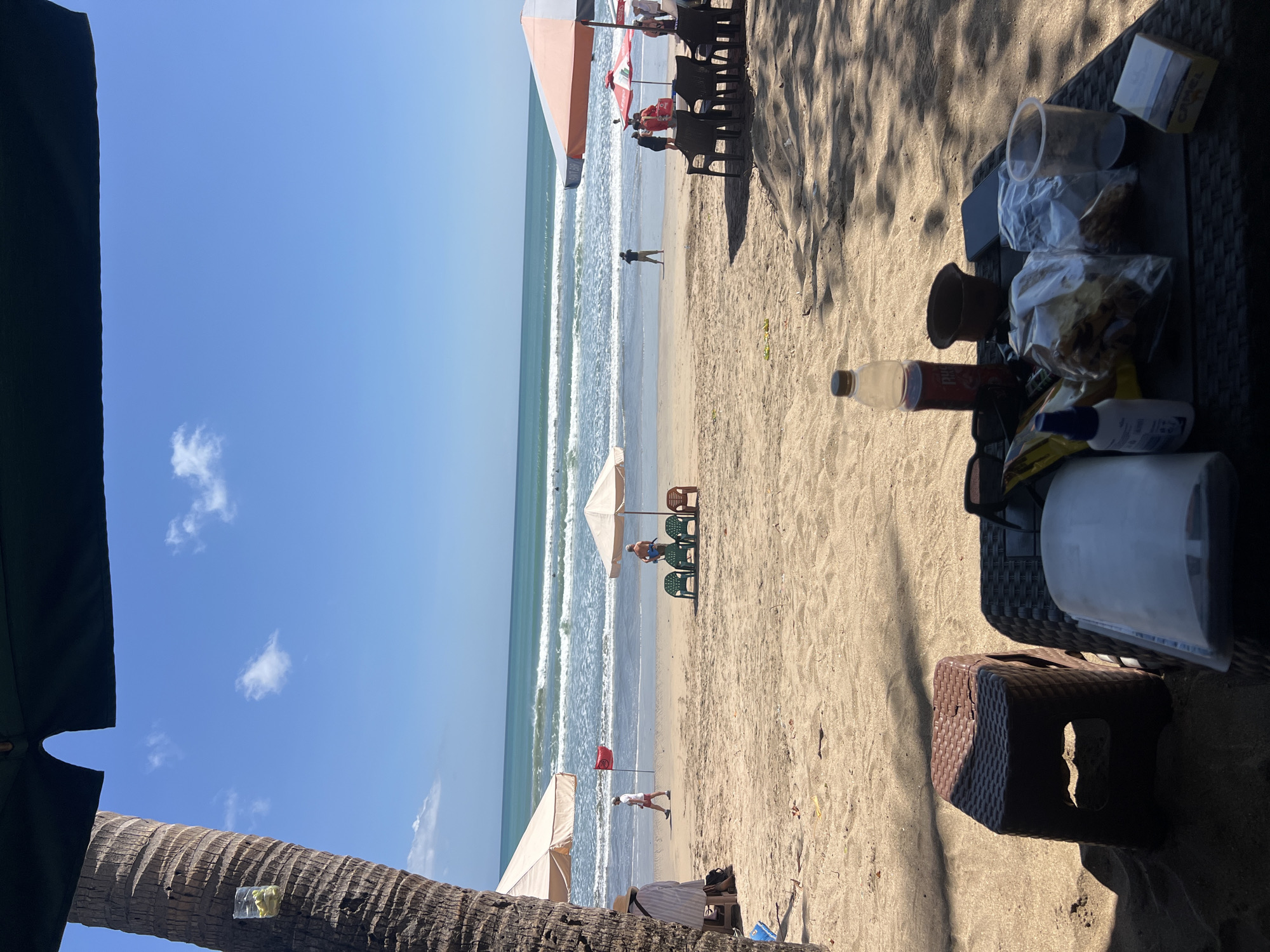 In Kuta, Indonesia, a sun-drenched beach scene unfolds under a bright blue sky dotted with scattered white clouds. The foreground is occupied by a low, dark-colored table laden with items. These include a clear plastic bottle with a dark liquid and a yellow cap, a small brown clay pot, a dark smartphone, two plastic bags containing food items, a clear plastic cup, a yellow and white box of what appears to be cigarettes, and a white bottle with a blue cap. Adjacent to the table is a small brown plastic stool. The table and stool are positioned on the sand, partially shaded by palm fronds.
Further out, the sandy beach stretches towards the ocean. Several beach umbrellas are visible, casting shadows on the sand. Some are white and peach, while others are beige. In the middle ground, a red flag is planted in the sand, and a man in a white shirt and red shorts walks along the shoreline. A group of four green plastic chairs and one brown plastic chair are set up near one of the umbrellas. Further offshore, several people are visible, some appearing to be swimming or surfing in the gentle waves. The ocean itself is a vibrant turquoise, meeting the horizon with a clear line. To the left, the textured bark of a palm tree trunk is prominent, with a small clear plastic bag filled with yellow objects tied to it.