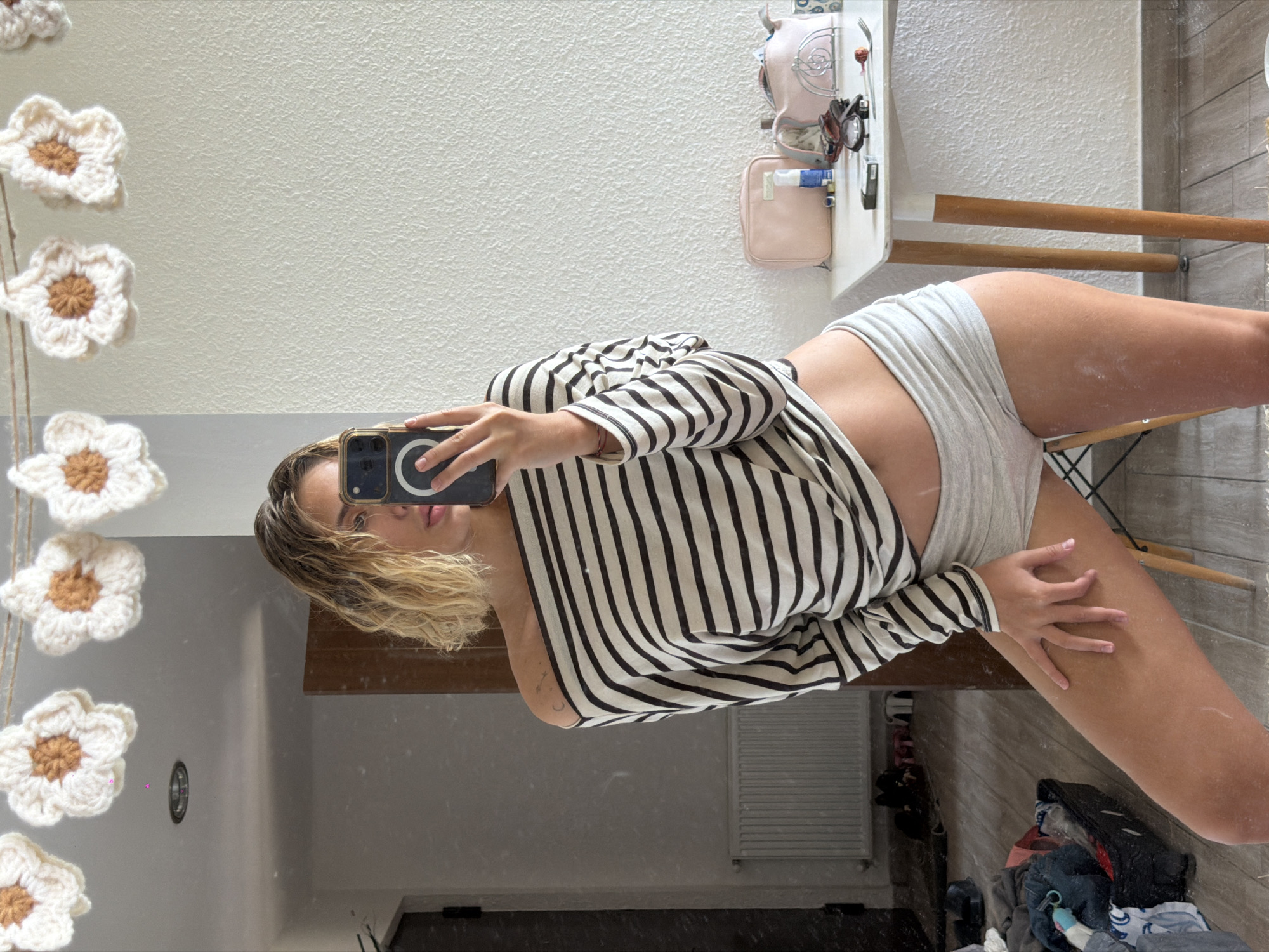 A woman with blonde, wavy hair is taking a mirror selfie in what appears to be an indoor domestic setting, possibly a bedroom or dressing area. She is wearing a black and white striped off-the-shoulder top and light grey shorts. She is holding a smartphone to take the photo, with her left hand resting on her thigh. In the background, to her left, a radiator is visible on a wall, and a doorway leads to another room. To her right, a white table holds several items, including makeup bags, a bottle, sunglasses, and a fork with a lollipop. A garland of crocheted white flowers with brown centers hangs across the top of the mirror. The floor appears to be wooden, with some items scattered around, including a black basket filled with clothes. The overall lighting suggests it is daytime. The location is not specified in the image or its details as being in Tbilisi, Georgia.