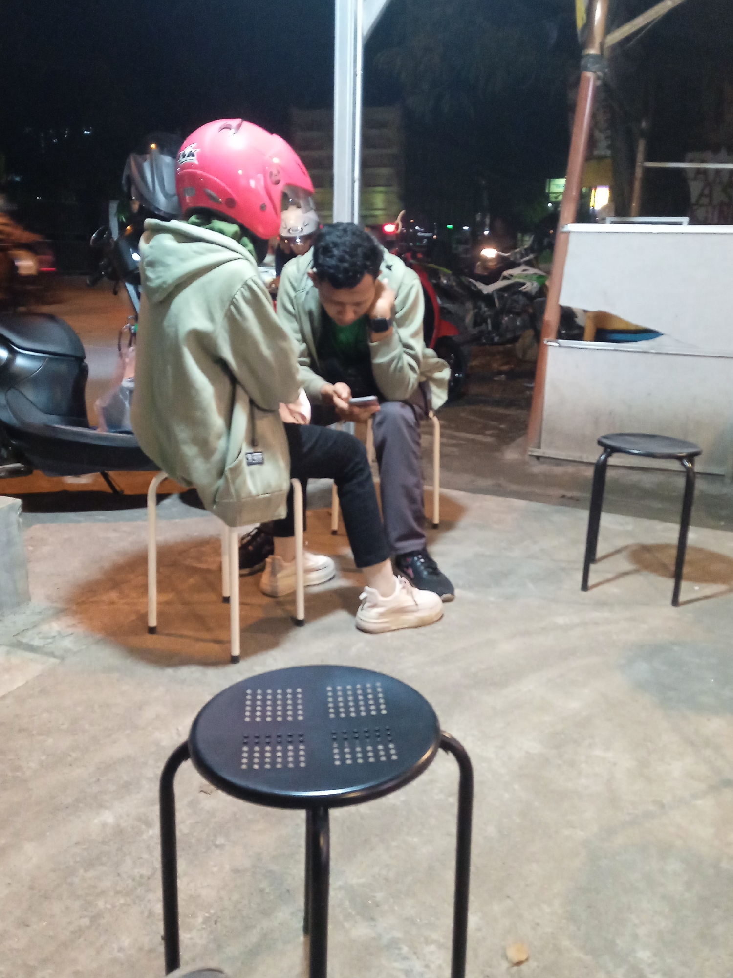 This image captures two individuals seated on simple metal stools at what appears to be an outdoor or semi-outdoor casual gathering spot at night in Sekopek Wetan, Indonesia. The ground is concrete, and artificial lighting illuminates the scene, casting shadows.
The person on the left is seen from behind, wearing a prominent pink motorcycle helmet with "INK" visible on the side. They are dressed in a light green or khaki hoodie over a green inner layer, black trousers, and white sneakers. To their left, a black scooter is partially visible.
The second individual sits facing partly towards the camera, looking down at a smartphone held in their hands. They are also wearing a similar light green or khaki hoodie over a green top, dark trousers, and black shoes. Their expression is focused on the phone.
In the immediate foreground, a black metal stool with a perforated circular top is positioned, acting as a table or an unoccupied seat. Another black stool stands unoccupied in the mid-ground on the right. In the background, several motorcycles are parked, and faint lights from other establishments or vehicles are visible in the dark surroundings. The overall setting suggests a relaxed, informal social interaction during the evening.