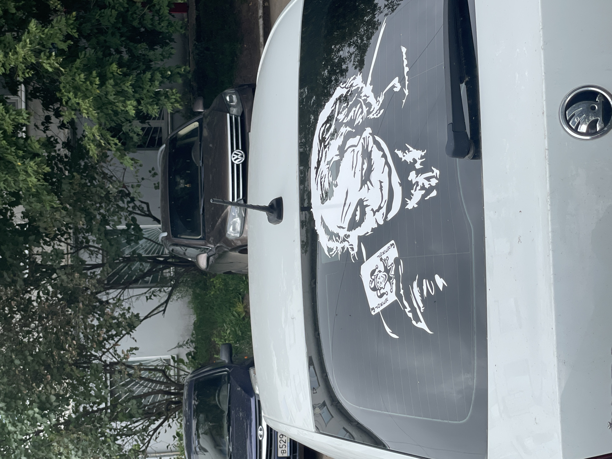 A white car's rear windshield is adorned with a large, white sticker of the Joker character, holding a playing card. The car is parked on an asphalt surface in front of a building with gray walls and windows, partially obscured by green trees and bushes. In the background, a dark-colored Volkswagen is visible, also parked. Another car, dark blue with a license plate starting with "B529", is parked to the left. The scene appears to be in a residential area, possibly a parking lot in the city of Chaikovsky, Russia. The weather seems overcast.