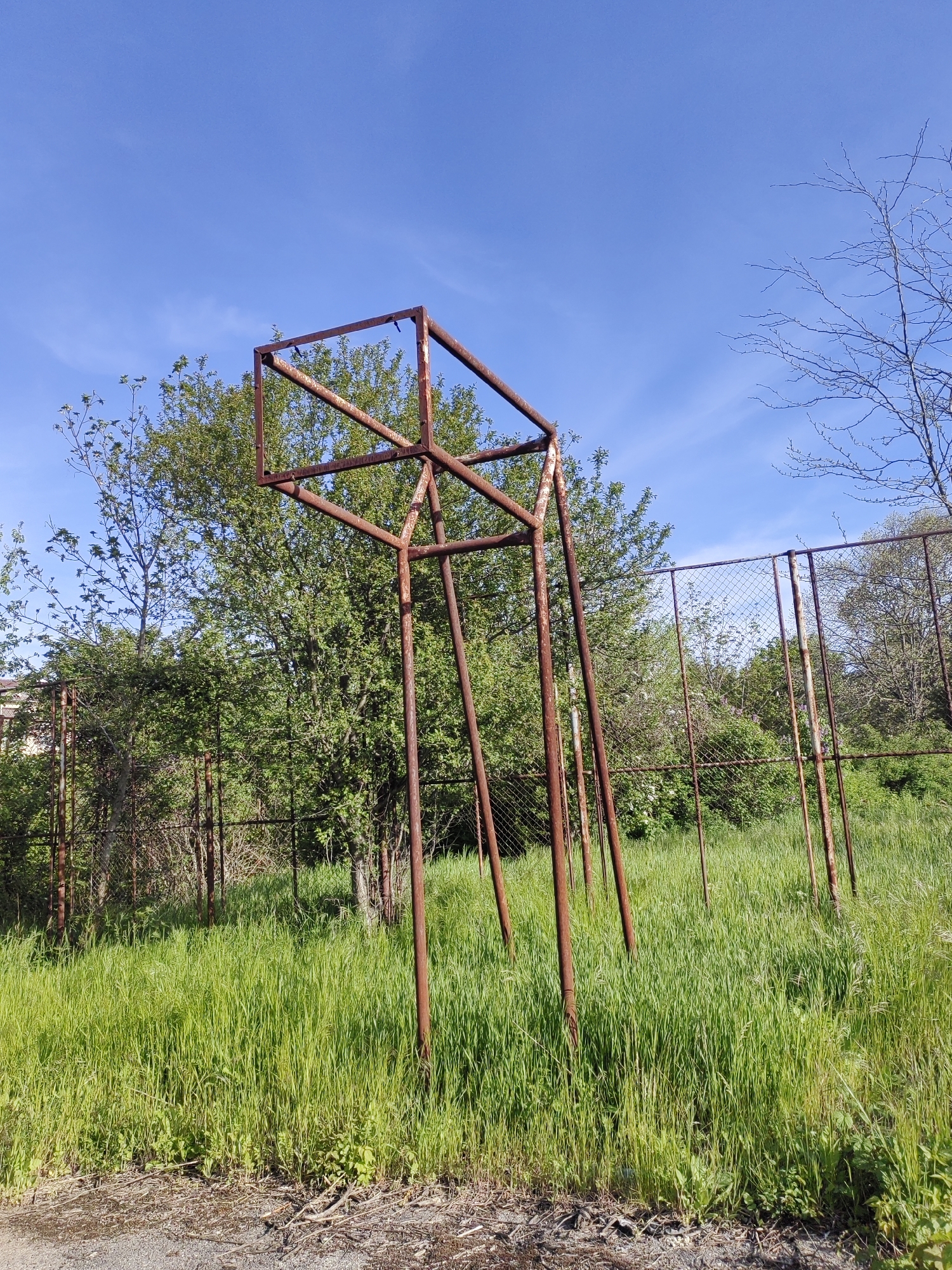 The image shows a rusted metal basketball hoop frame without a backboard or net, standing in a grassy field in what is potentially an abandoned or neglected area. The location is suggested to be Dobrich, Bulgaria. The hoop is constructed of rusted metal poles forming a square backboard frame and four supporting legs. Tall green grass surrounds the base of the hoop. A rusted chain-link fence runs along the perimeter behind the basketball hoop. Trees and other foliage are visible behind the fence. The sky is blue with light clouds. The ground near the bottom of the image is a mix of dirt and gravel. No people are visible in the image.