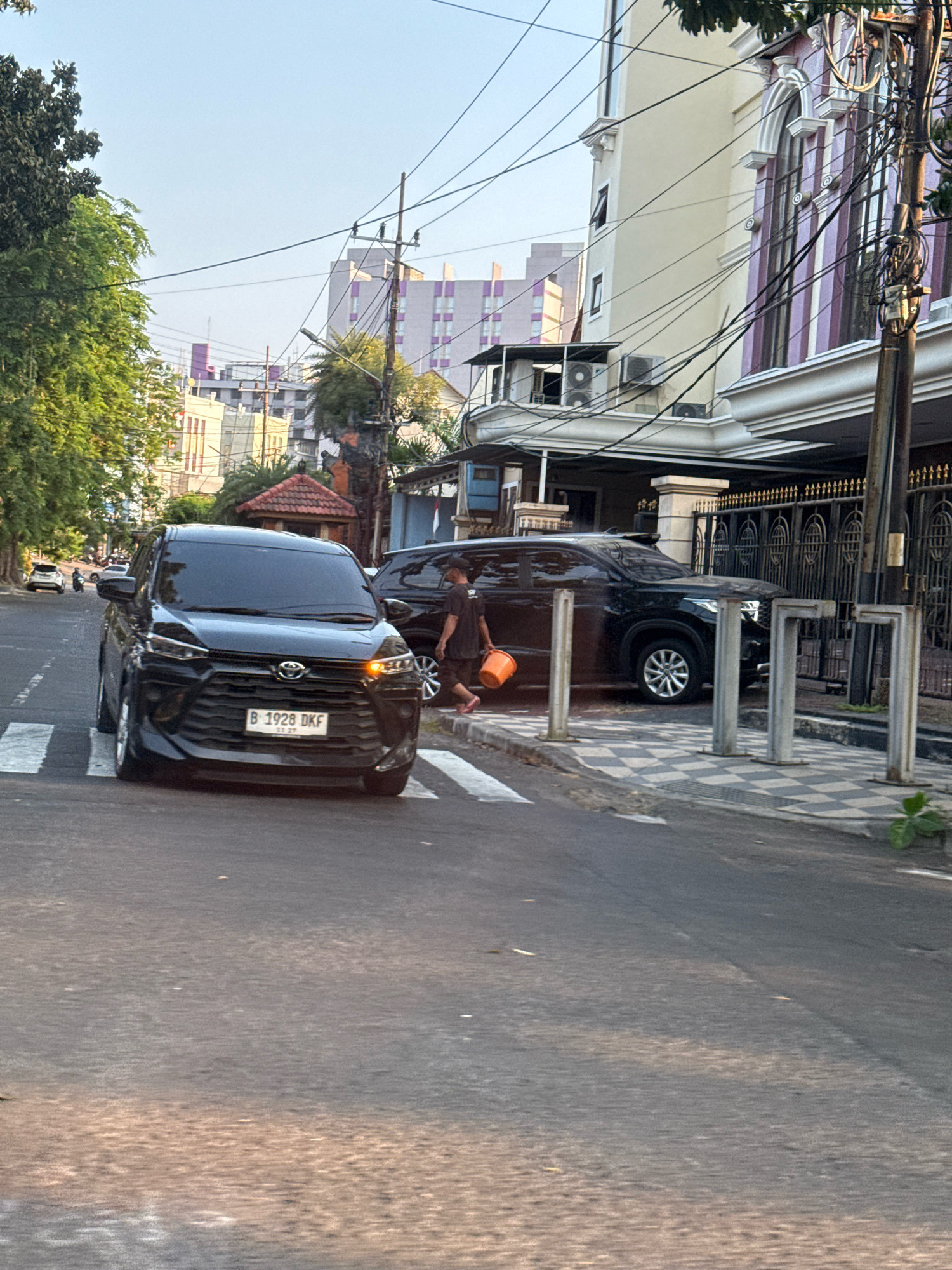 The image depicts a street scene in Surabaya, Indonesia, with a black SUV prominently in the foreground crossing a pedestrian crosswalk. The SUV has a license plate that reads "B 1928 DKF." To the right of the SUV, a person, possibly male, is walking away from the vehicle carrying an orange bucket. Behind the black SUV, another dark-colored SUV is parked. The street is paved, and there are buildings lining the right side of the road, one of which has ornate architectural details and decorative pillars. The environment appears to be urban, with trees on the left side of the street and utility poles with numerous wires overhead. The lighting suggests it is either early morning or late afternoon, with the sun casting long shadows. The background shows more buildings, some of which are tall and modern, indicative of a city setting. The scene is captured from a slightly elevated perspective, looking down at the street.