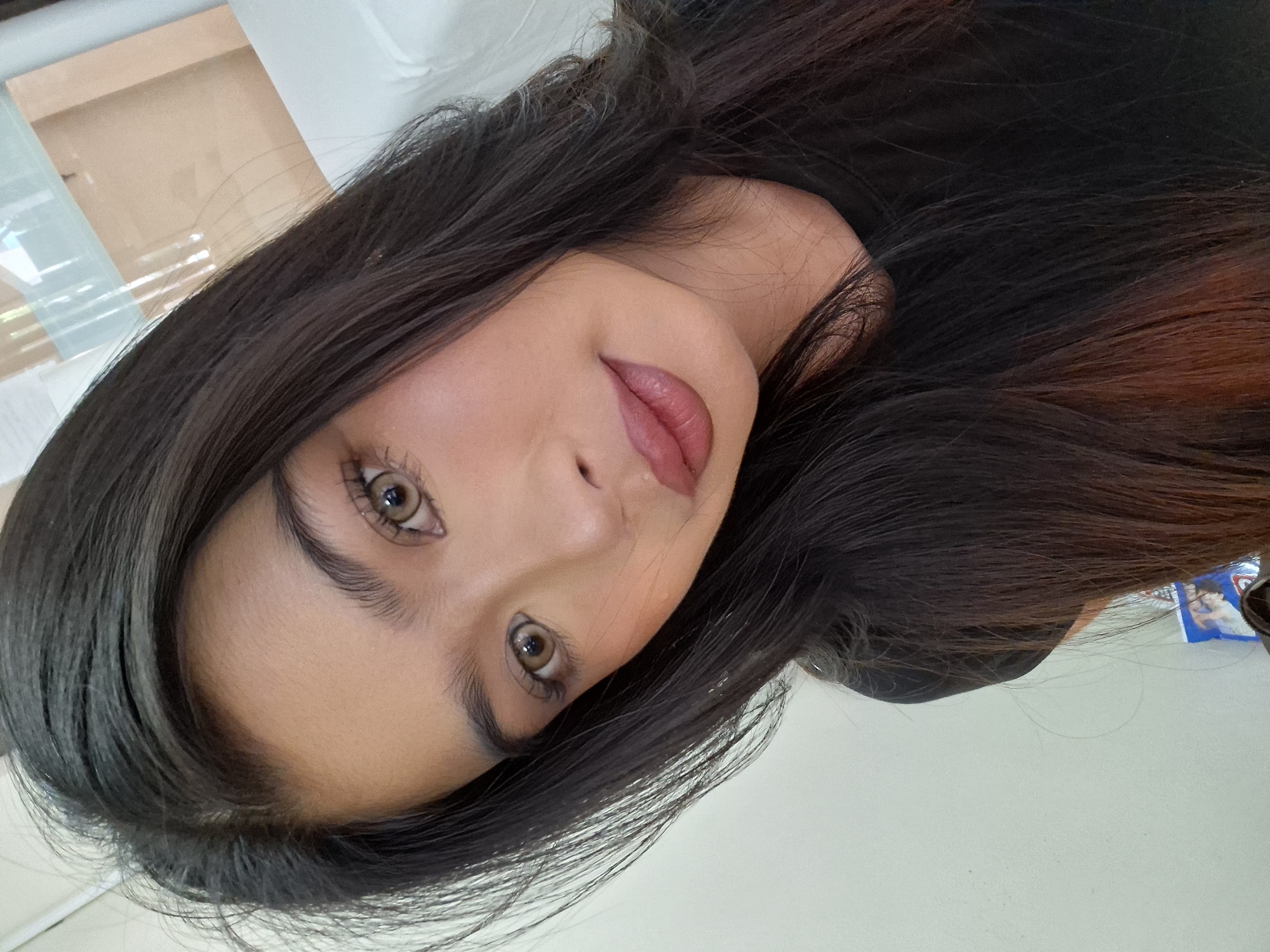 A close-up, slightly angled selfie shows a young woman with long, dark hair framing her face. She is looking directly at the camera, her head tilted slightly to the left. Her makeup is noticeable, with defined eyebrows, subtle eyeshadow, mascara, and a matte, rosy-toned lipstick. She is wearing a black t-shirt, and her skin appears smooth with a warm undertone. The background is softly blurred, suggesting an indoor environment with a light-colored wall and a hint of a reflective surface, possibly a window or a screen, behind her to the right. There are no discernible landmarks or elements that would place this image specifically in Kuta, Indonesia.