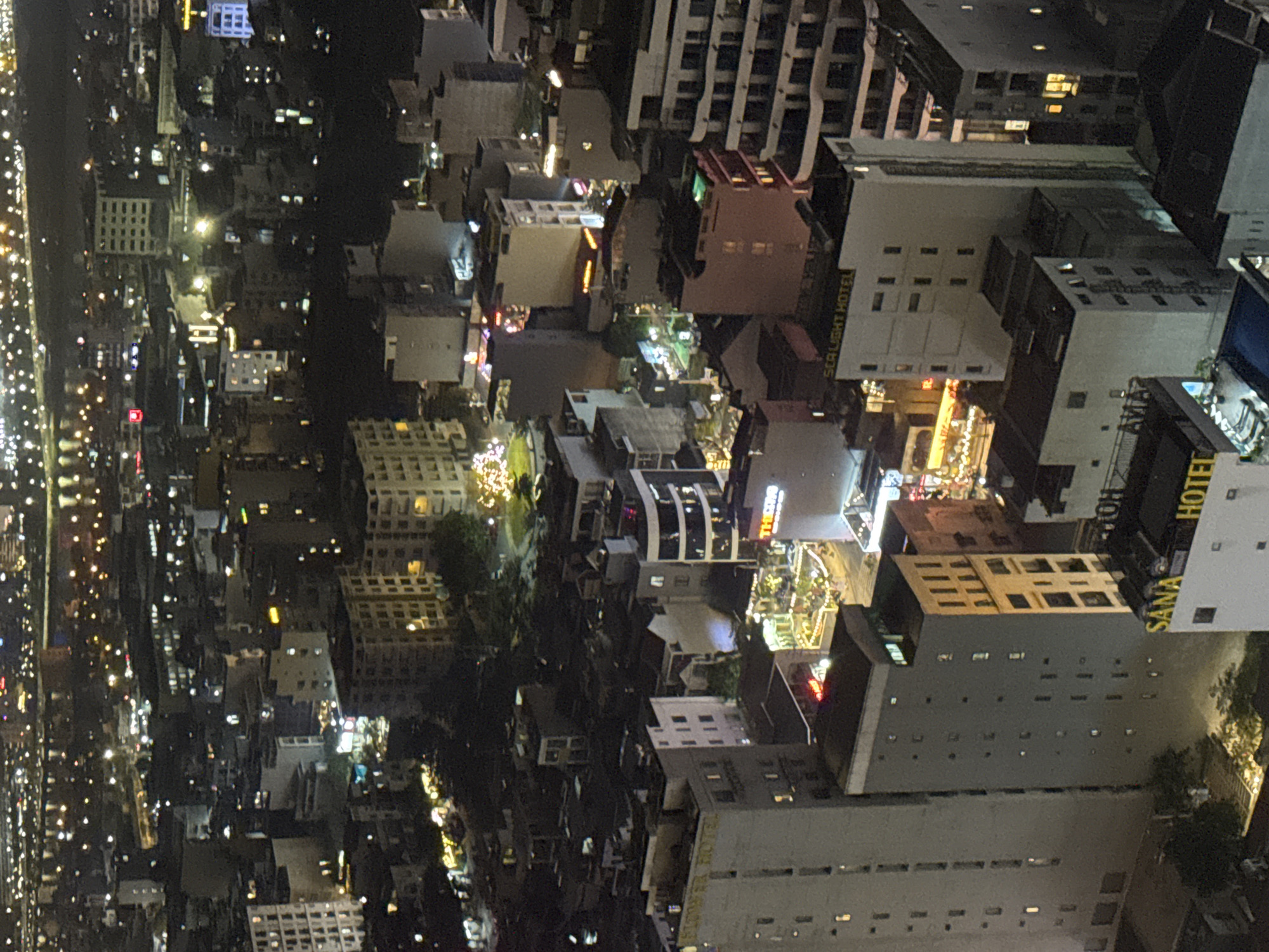 The image captures an aerial nighttime view of Đà Nẵng, Vietnam, dominated by illuminated buildings and streets. The cityscape is dense with high-rise structures and residential blocks. Streetlights and building lights create a grid pattern, highlighting the city's layout. The Sana Hotel is also visible within the shot.