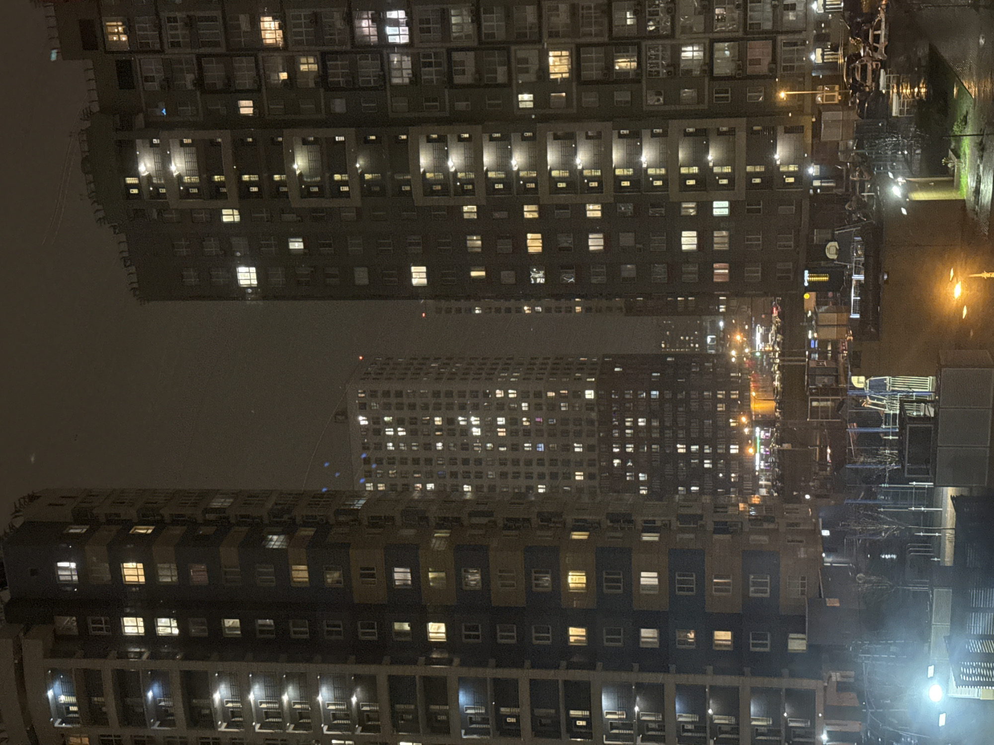 The image depicts a nighttime scene in Khimki, Russia, showcasing dense residential buildings with numerous illuminated windows. Two tall apartment buildings flank a third, more distant building in the center. The right side of the image shows streetlights reflecting on wet surfaces, indicating possible rain. The general atmosphere suggests a densely populated urban area at night, with signs of human activity through lighted windows. Construction materials, possibly for building maintenance, appear on the right side of the photo near the apartments.