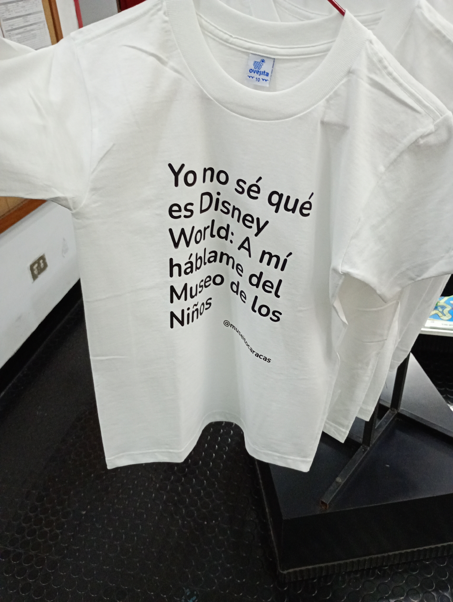 A white short-sleeved t-shirt with a round neck is displayed indoors, likely in a store or exhibition space. The t-shirt is the primary focus, presented hanging on a rack or stand, with other similar t-shirts partially visible in the background. The floor beneath is black with a textured, circular pattern.
The main text printed on the t-shirt in black, sans-serif font reads: "Yo no sé qué es Disney World: A mí háblame del Museo de los Niños". This translates to "I don't know what Disney World is: Talk to me about the Children's Museum." Below this, in smaller font and curved, is the social media handle "@museitocaracas". This handle strongly suggests a connection to the Museo de los Niños de Caracas, aligning with the provided location context of Caracas, Venezuela.
The t-shirt's brand tag, visible at the back of the neck, shows a small logo of a sheep and the word "ovejita" (little sheep), along with "Talla 10" (Size 10), indicating it might be a child's or youth's size. The overall impression is a statement piece promoting a local cultural institution over a globally recognized theme park.