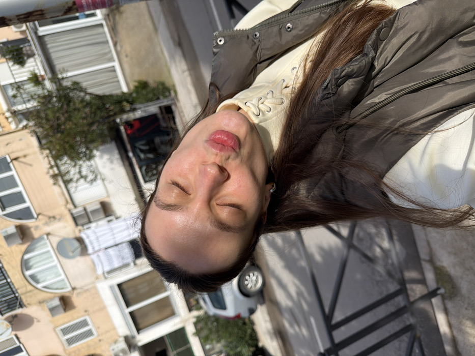 A female subject is depicted in a close-up, angled outdoor selfie, her head tilted to the viewer's left. Her eyes are closed, and her lips are pursed in a kissing gesture. She has long, dark brown hair and wears a light-colored, high-collared top with a laced front, beneath a dark olive green puffer vest. A small silver hoop earring is visible in her left ear.
The scene is an urban street in Budva, Montenegro, under bright sunlight, which casts sharp shadows. In the background, a multi-story building with a light beige facade features various windows, some with external shutters, and two arched windows. Multiple air conditioning units and two satellite dishes are mounted on the building's exterior. White laundry is visible hanging to dry from an upper-floor window. A paved street or road is present, partially obscured by a dark metal railing in the foreground. A silver station wagon is parked or moving on the street to the left, and a darker vehicle is further down the street. Sparse trees are visible between the street and the buildings. A utility pole on the right side of the frame displays a red and white sign containing text, including the word "MONTENEGRO," confirming the country. The environment is consistent with a residential or mixed-use urban street.