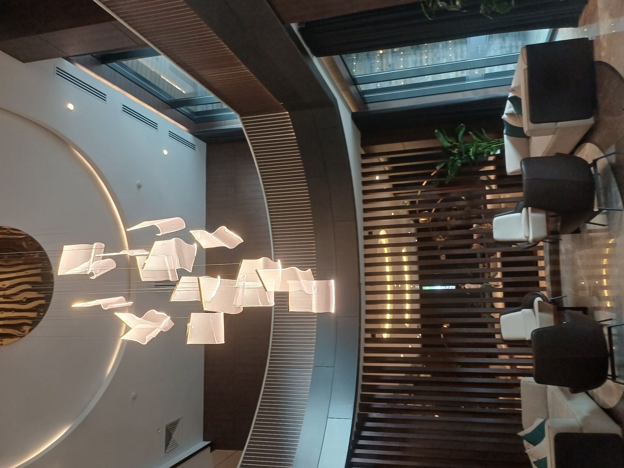 The image depicts the interior of a modern, upscale lobby or waiting area, likely in a hotel or commercial building. The scene is dominated by a large, multi-tiered contemporary chandelier featuring numerous translucent, cube-like elements suspended by thin wires from a circular, metallic ceiling fixture.
The architecture of the space is characterized by curved walls and multiple levels, indicated by a balcony with a white railing visible in the mid-ground. The walls are finished with dark wood paneling and contrasting dark brown wall coverings. The flooring appears to be polished marble or a similar reflective material.
Furnishings include several comfortable seating arrangements. On the left, a light-colored sectional sofa with turquoise accent pillows is partially visible. In the foreground, a dark charcoal armchair with a metallic base is prominently featured. To the right of this, a lighter-colored armchair with a dark base is positioned near a small table. Further back, another light-colored sofa with dark green pillows is situated, accompanied by large potted plants that add a touch of greenery to the sophisticated ambiance.
Large windows in the background reveal a glimpse of the exterior, possibly showing other buildings or city lights, suggesting an urban setting. The overall impression is one of contemporary elegance and spaciousness.
The location in the city of Myk, Uzbekistan is not discernible from the visual evidence provided in the image.