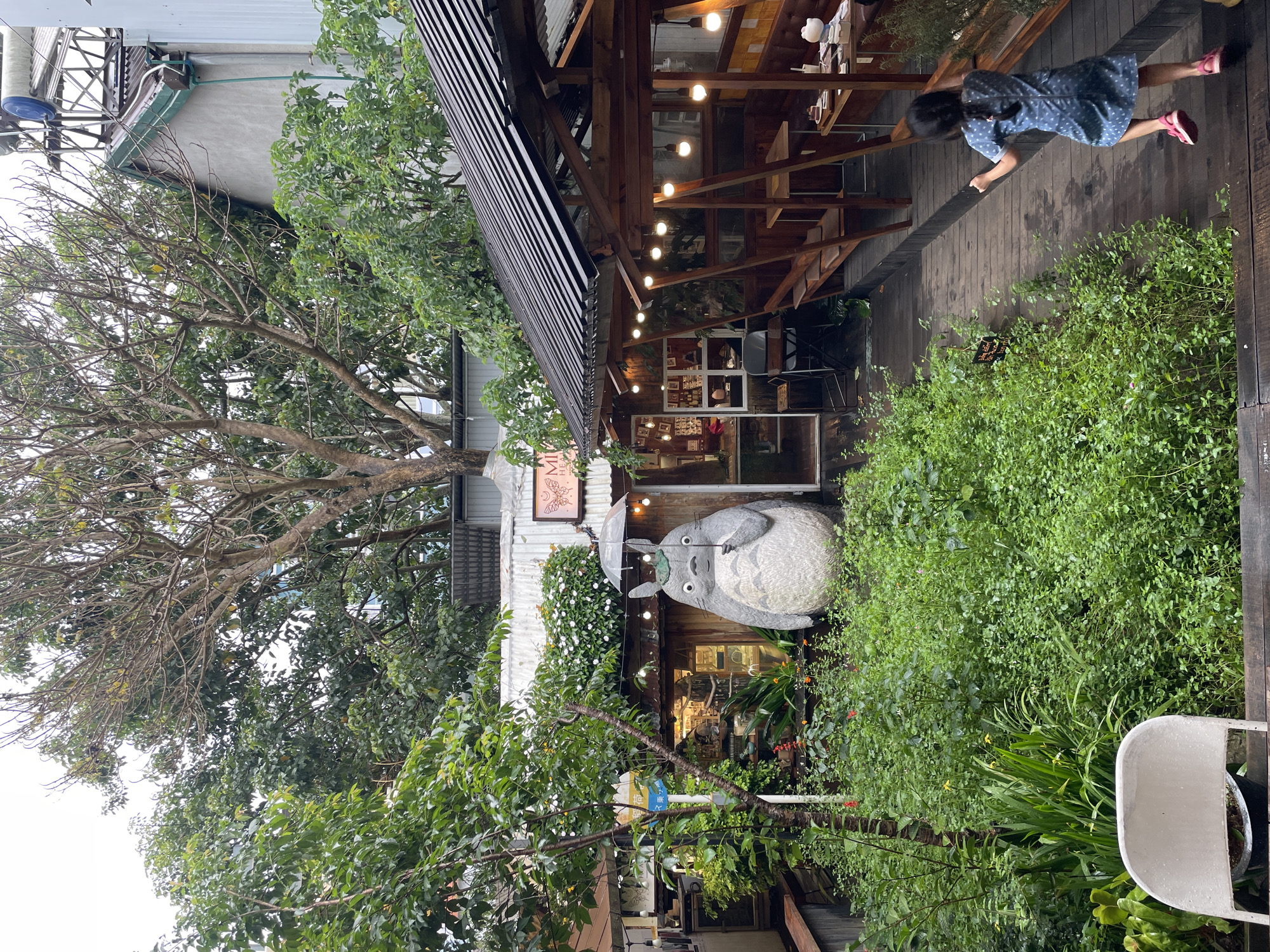 A view of a café with a statue of Totoro holding an umbrella outside the entrance. The café has a wooden deck and roof. Green vegetation is abundant around the café and there is a large tree to the left. A girl is running on the deck. The café appears to be in Lâm Viên - Đà Lạt, Vietnam.