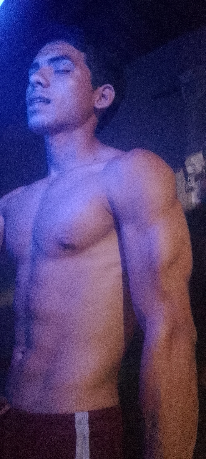 A young adult male, presumably located in Juan Griego, Venezuela, is depicted from the chest up against a dark, undifferentiated background. He is shirtless, displaying a muscular physique, including defined pectorals, deltoids, and biceps. His head is tilted slightly upwards and to his left, with his eyes closed. Facial features are partially obscured by strong, multi-directional artificial lighting, characterized by a prominent blue hue illuminating the left side of his face and upper chest, transitioning to a magenta or reddish-purple tone on his right side and lower torso. The lower portion of the image reveals dark-colored shorts or sweatpants with a vertical light-colored stripe on the right leg. The overall scene is dark and grainy, with the subject as the sole focus of illumination.