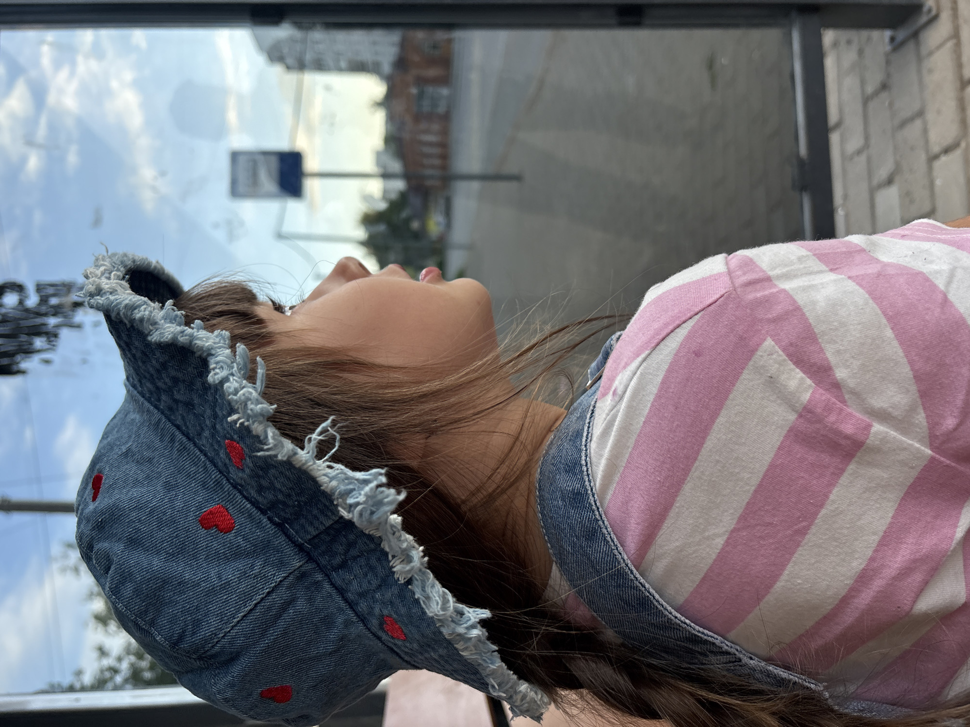 A young individual, depicted from a right-side, slightly rear perspective, gazes outwards through a large glass window onto a city street in Chaikovsky, Russia. The subject wears a denim bucket hat adorned with multiple small red embroidered hearts and featuring a visibly distressed, frayed brim. Light brown hair is visible beneath the hat, with a portion styled into a braid resting over the subject's right shoulder (viewer's left). The attire includes a white t-shirt with horizontal pink stripes and denim overalls, with one strap evident over the right shoulder. The subject's profile shows the eyes directed towards the outside scene, with lips slightly parted. Reflections on the glass window include blurred shapes and text-like patterns. Beyond the window, a paved sidewalk and street extend into the distance. A blue and white bus stop sign is centrally located in the mid-ground. In the background, various urban structures are visible, including an older brick building and multi-story residential buildings, beneath a sky with scattered clouds.