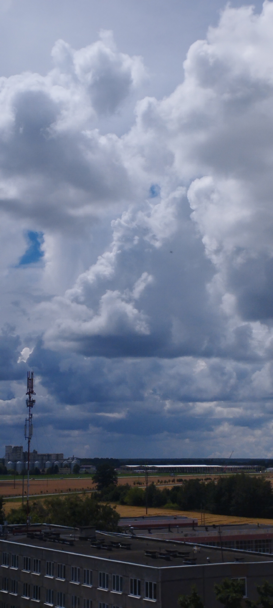This is a vertical, medium-shot, outdoor photograph taken from a high vantage point, looking slightly down at a landscape under a dramatic sky. The sky is filled with large, white, and grey cumulus clouds, with patches of blue sky visible. In the mid-ground, a cell tower stands prominently on the left, with multiple antennas. Behind the tower, several large, round silos are visible, along with a long, low-rise industrial building. Further in the distance, fields of yellow crops stretch towards a line of trees and other structures. The foreground of the image is dominated by the flat, dark-grey roof of a building, showing rows of evenly spaced vents and a section of the building's facade with multiple rectangular windows. The overall scene suggests an industrial or semi-urban area within Skidzieĺ, Belarus, on a partly cloudy day.