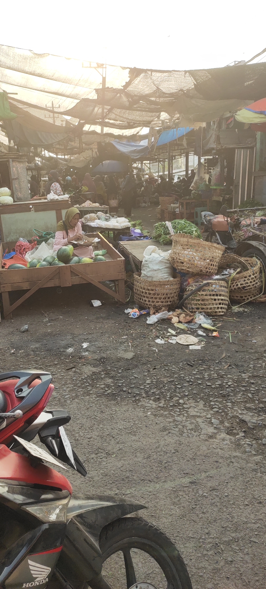 This image depicts a bustling open-air market, likely in Karangtowo, Indonesia, under a canopy of tarps and netting that filters the bright sunlight. The ground is a mix of dirt and gravel. In the foreground, the front portion of a red and black motorcycle is visible. The mid-ground features a woman in a pink hijab and dress sitting in a wooden stall filled with watermelons and other produce. She appears to be working, possibly preparing or arranging the fruit. Adjacent to her stall are various baskets, some woven and some made of fabric, filled with produce and what appear to be bags of goods. Some trash is scattered on the ground around the stalls. In the background, numerous people are browsing and shopping, with more stalls and structures indicating a well-established market environment. The overall scene is one of everyday commerce and activity in a vibrant local market.