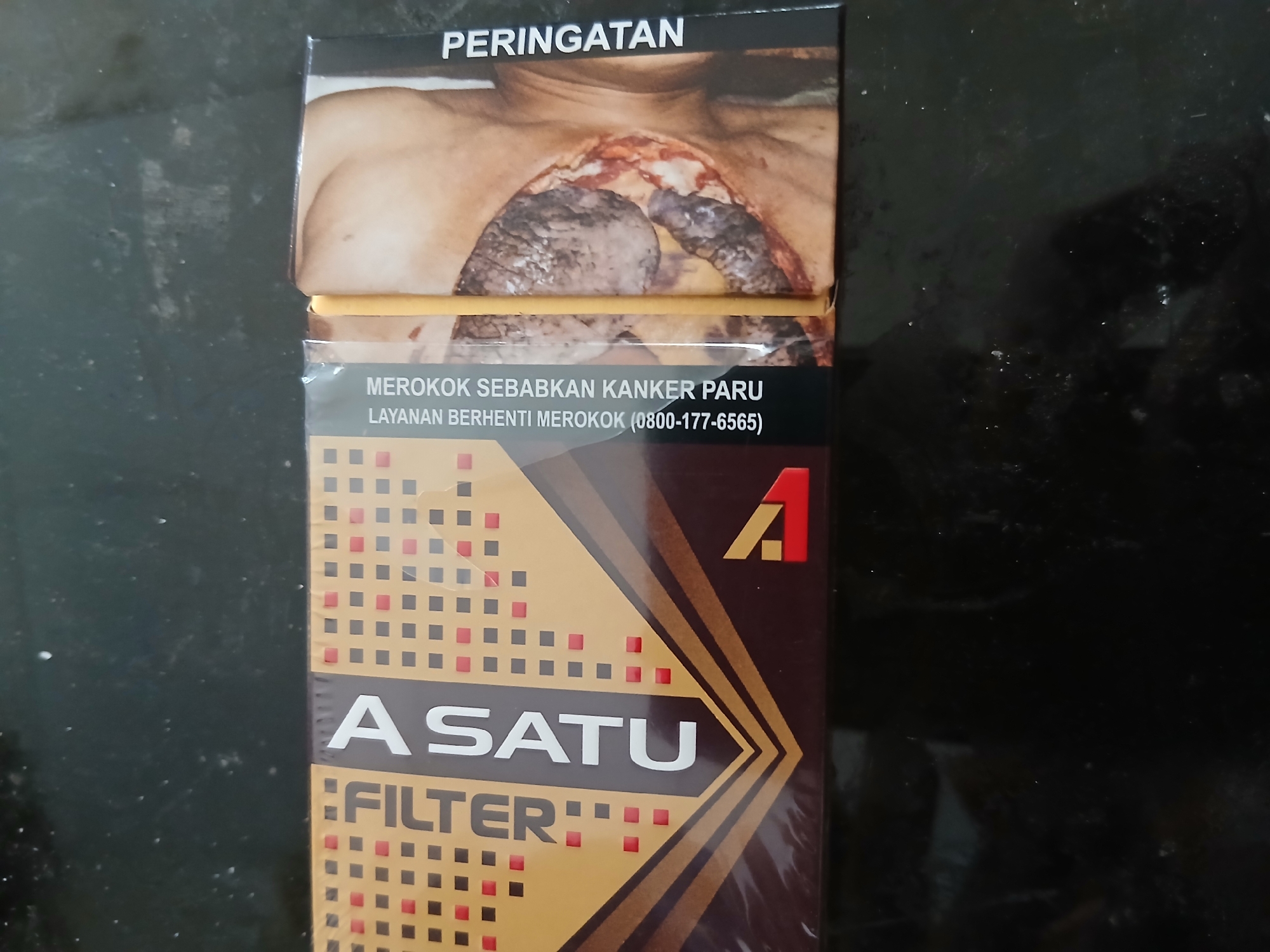 A close-up, high-angle shot captures a pack of ASATU Filter cigarettes resting on a dark, speckled surface, likely a table. The cigarette pack is oriented horizontally, with its front face prominently displayed. The pack features a design with a dark brown base and a patterned area in shades of gold and brown squares. The brand name "ASATU" is emblazoned in large, white, sans-serif lettering, with "FILTER" written vertically in smaller white text to its left. A graphic of a human torso with a severe medical condition, displaying exposed diseased tissue, is visible on the right side of the pack, accompanied by the Indonesian text "PERINGATAN MEROKOK SEBABKAN KANKER PARU LAYANAN BERHENTI MEROKOK (0800-177-6565)". The image does not contain any people or provide information about its location in Tenggarang, Indonesia.