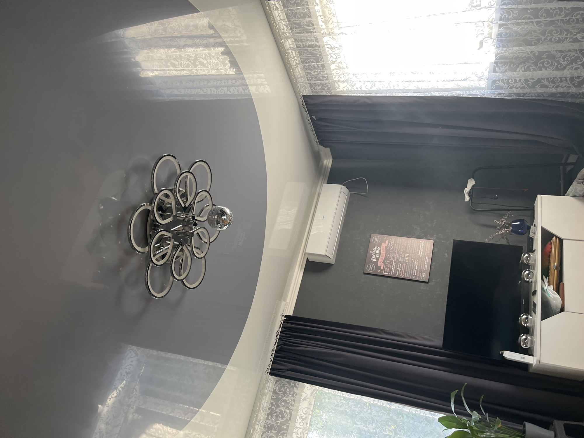 This is a photograph of an interior room, likely a living room, with a view towards a window. The room features a modern, multi-ringed ceiling light fixture with a disco ball hanging from its center. The ceiling itself is a glossy, dark gray stretch ceiling, reflecting the light from the window and the fixture.
On the wall, there is a dark gray textured finish. Mounted on this wall is a white air conditioning unit. To the left of the air conditioner, a black curtain is drawn, partially obscuring a window with sheer white patterned curtains. To the right of the air conditioner, another dark curtain is drawn, also alongside a window with sheer white patterned curtains.
Below the air conditioner and between the two sets of curtains, a black television is placed on a white media console. The media console has shelves, one of which contains various items, including what appears to be a wooden box and some white plastic bags. In front of the television, on the console, are four spherical decorative objects. To the left of the television, a green potted plant is visible. On the right side of the console, a blue vase with dried flowers is present.
A poster with text in Cyrillic script is mounted on the wall above the media console, between the air conditioner and the right curtain. A tall, dark metal stand with some items hanging from it is positioned to the right of the media console, partially obscured by the dark curtain.
The lighting in the room appears to be a mix of natural light coming through the windows and artificial light from the ceiling fixture. The overall impression is of a modern, somewhat dark-toned living space. The photograph was taken in Saratov, Russia.
