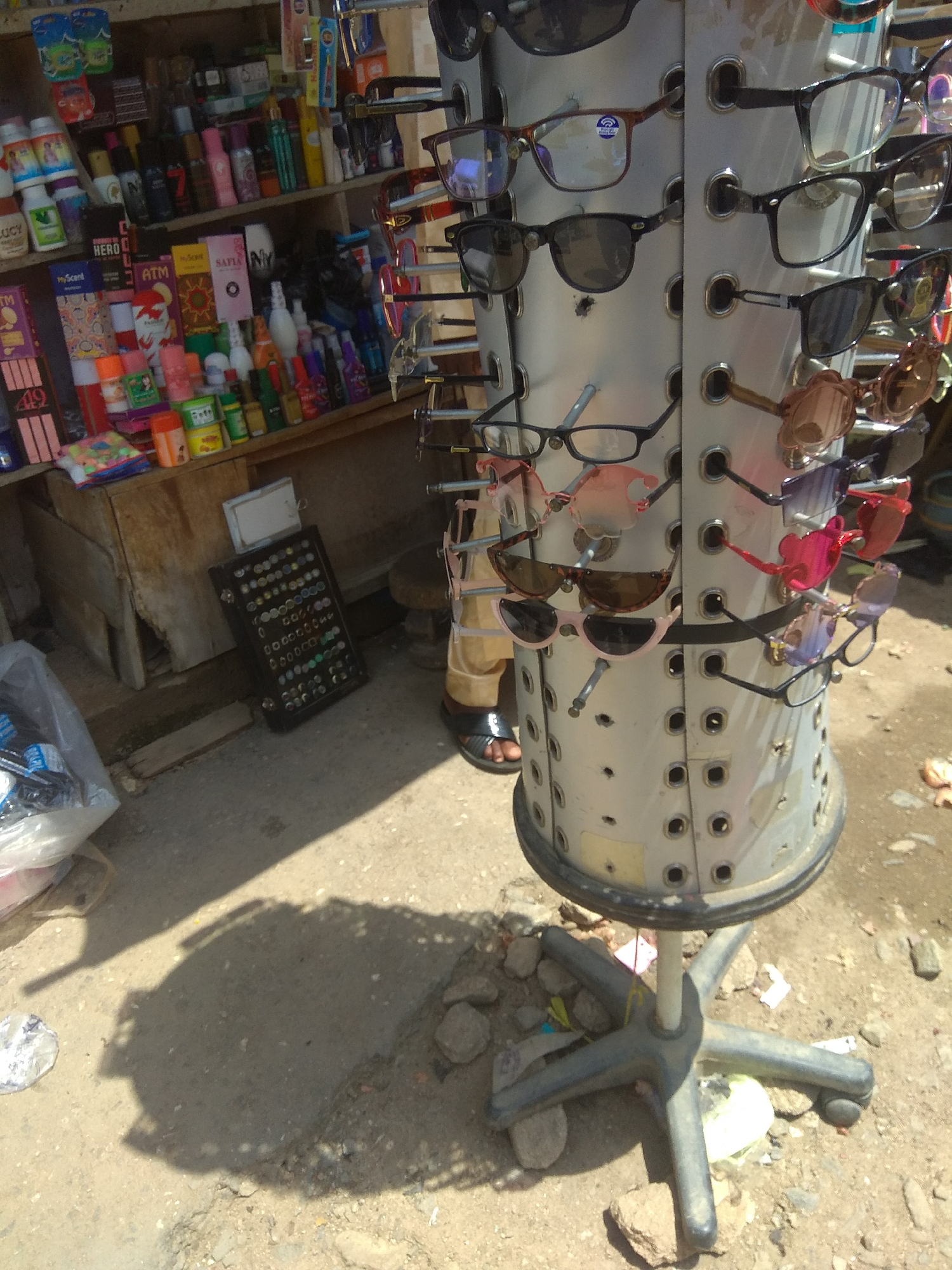 The image shows a street vendor's stall in Jalingo, Nigeria, with a rotating display of sunglasses. The stall is set up outdoors, with a focus on the display rack of eyewear. The rack is a cylindrical, metallic structure with multiple levels, each holding several pairs of sunglasses. The sunglasses vary in style, color, and frame type, suggesting a variety of options for potential customers. Behind the sunglasses display, shelves are visible, stocked with various consumer goods such as bottles of liquids, boxed items, and what appear to be personal care products. The ground is unpaved, with sand, gravel, and some scattered debris, typical of a market environment in Nigeria. A shadow cast by the display rack suggests a sunny day. The presence of the vendor's feet in sandals at the bottom right indicates a person operating the stall, though no other individuals are clearly visible. The scene depicts a commercial activity within the urban landscape of Jalingo.
