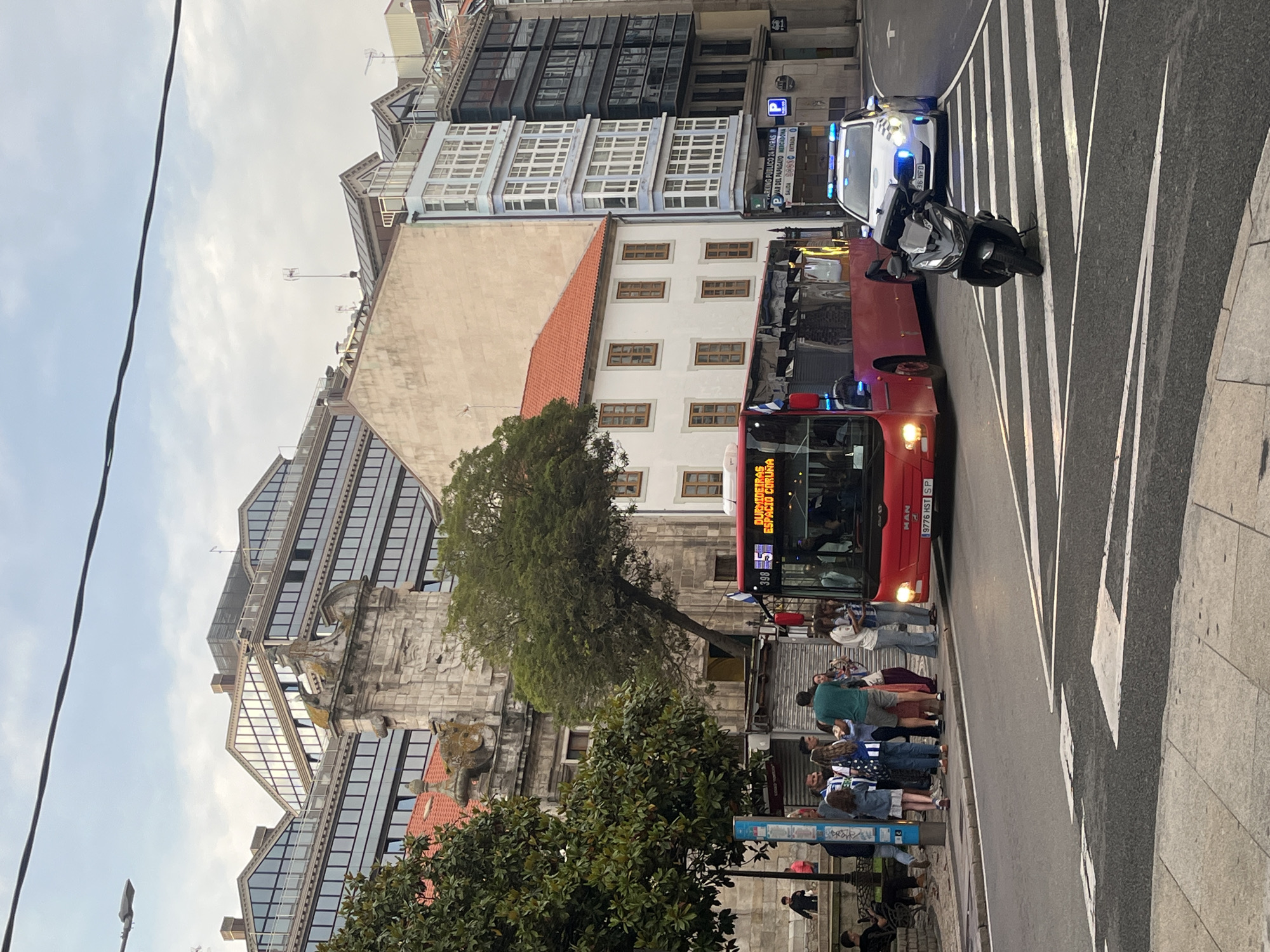The image captures a street scene in A Coruña, Spain. A red city bus is stopped on the road, its route number displaying "5" and destination reading "DURMIDEIRAS ESPACIO CORUÑA". A group of people is standing on the sidewalk near the bus, some of whom are wearing blue and white Deportivo de La Coruña jerseys. A black scooter and a police car with its lights on are parked behind the bus on a zebra crossing. Buildings of varying architectural styles line the street, with one featuring a stone facade and another displaying a "P" parking sign. An overhead power line is visible in the upper left corner.