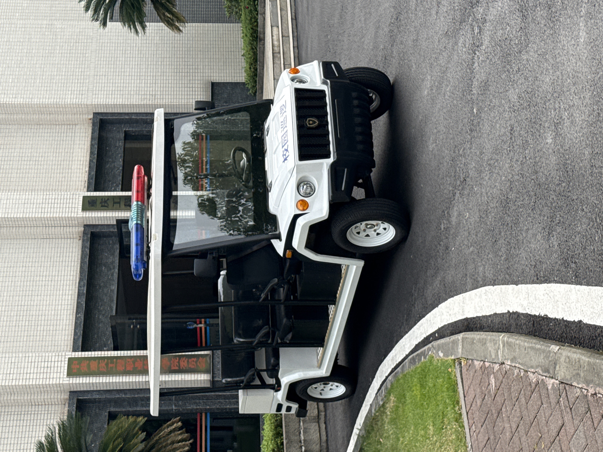 A white, open-air electric vehicle, resembling a golf cart with a roof and seating for multiple passengers, is parked on an asphalt road. The vehicle has a blue and red light bar mounted on its roof and Chinese characters on its front windshield. The background features a building with a tiled facade and palm trees. A brick-paved walkway and a grassy area are visible to the left of the vehicle. The vehicle is positioned in front of a building with a sign that reads "重庆工程职业技术学院" (Chongqing Vocational College of Engineering). The location is the campus of Chongqing Vocational College of Engineering in Jiangjin District, Chongqing, China. The scene is static, with no individuals or ongoing actions depicted.