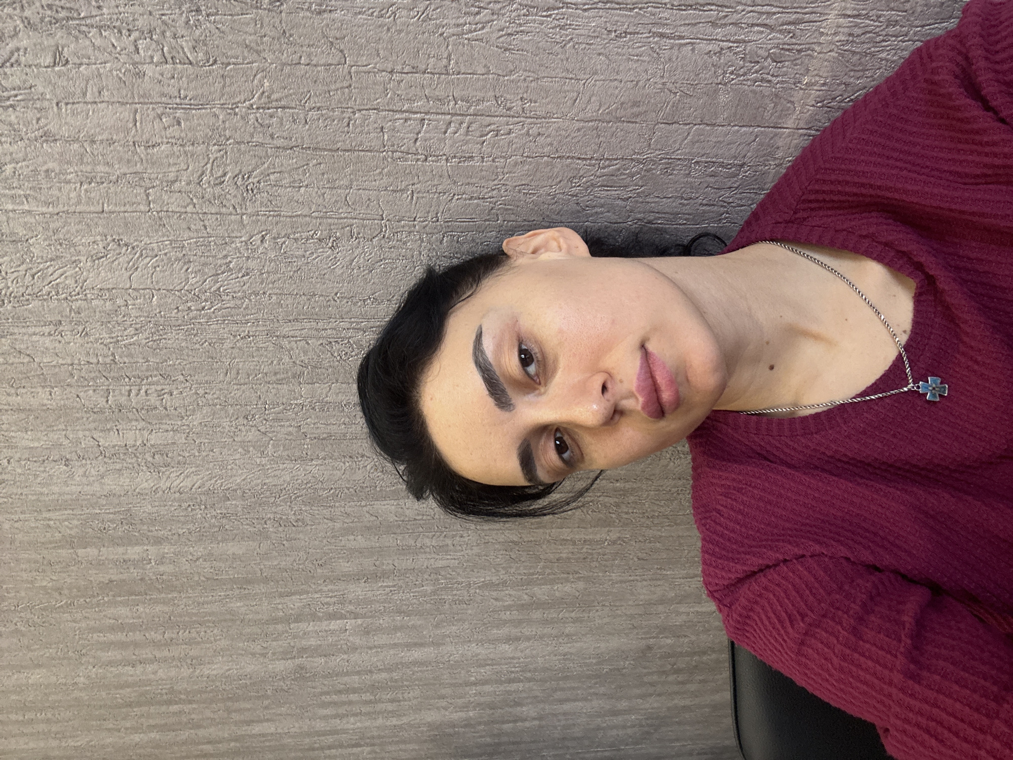 An adult female is depicted in a head-and-shoulders portrait, positioned slightly right of center and facing directly forward. She has dark hair, dark eyes, and full lips, with visibly enhanced eyebrows. She is wearing a dark red, textured long-sleeved garment and a silver-colored chain necklace with a small, light blue cross pendant. A dark, possibly leather or synthetic, chair back is visible in the lower left portion of the frame. The background consists of an indoor wall with a muted grey-brown, vertically textured pattern. There are no discernible features to indicate a specific location within the city of Tbilisi, Georgia.